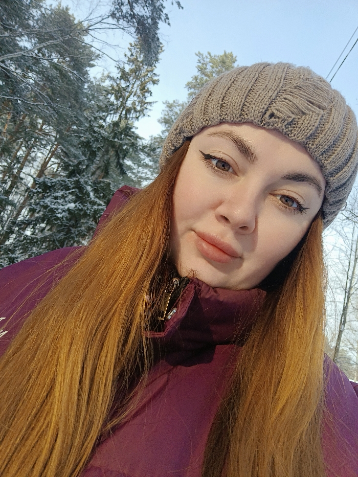 A close-up, selfie-style photograph depicts a young adult female positioned in the foreground against a snowy, wooded background. The female has long, reddish-blonde hair cascading over the shoulders of a plum-colored, high-collar puffer jacket. A gold-colored safety pin is secured to the inner flap of the jacket near the collar. She wears a light brown ribbed knit beanie, exhibiting small tears or wear on its top and left side. Her face features fair skin, light brown eyes accentuated by winged dark eyeliner, and well-defined eyebrows. Her lips are full with a natural, light pink tone. She maintains direct eye contact with the camera, exhibiting a neutral to slightly smiling expression. The background consists of densely snow-covered coniferous trees and some bare deciduous trees under a clear, light blue sky, indicating a winter landscape. The specific location within Chaikovsky, Russia, cannot be precisely identified from the visual evidence, but the scene is consistent with a forested or park area during winter.