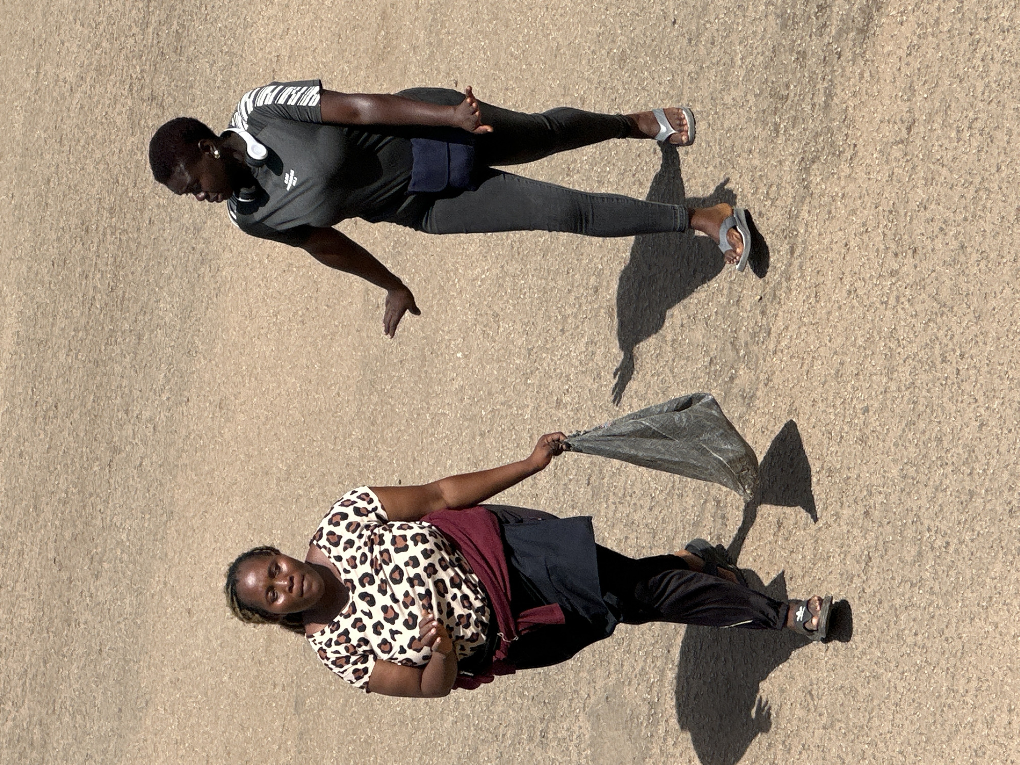 Two women are walking on a dirt road in Bukuru, Nigeria. The woman on the left is wearing a leopard-print t-shirt, a maroon sash tied around her waist, a dark skirt, and black pants. She is carrying a gray plastic bag in her right hand and is looking downwards. The woman on the right is wearing a dark gray t-shirt with white stripes on the sleeves, dark pants, and silver flip-flops. She has headphones around her neck and a small navy blue pouch attached to her waist. She is looking to her left and has her right hand extended outwards. The ground is composed of coarse, light brown dirt. The lighting suggests it is daytime, casting distinct shadows.