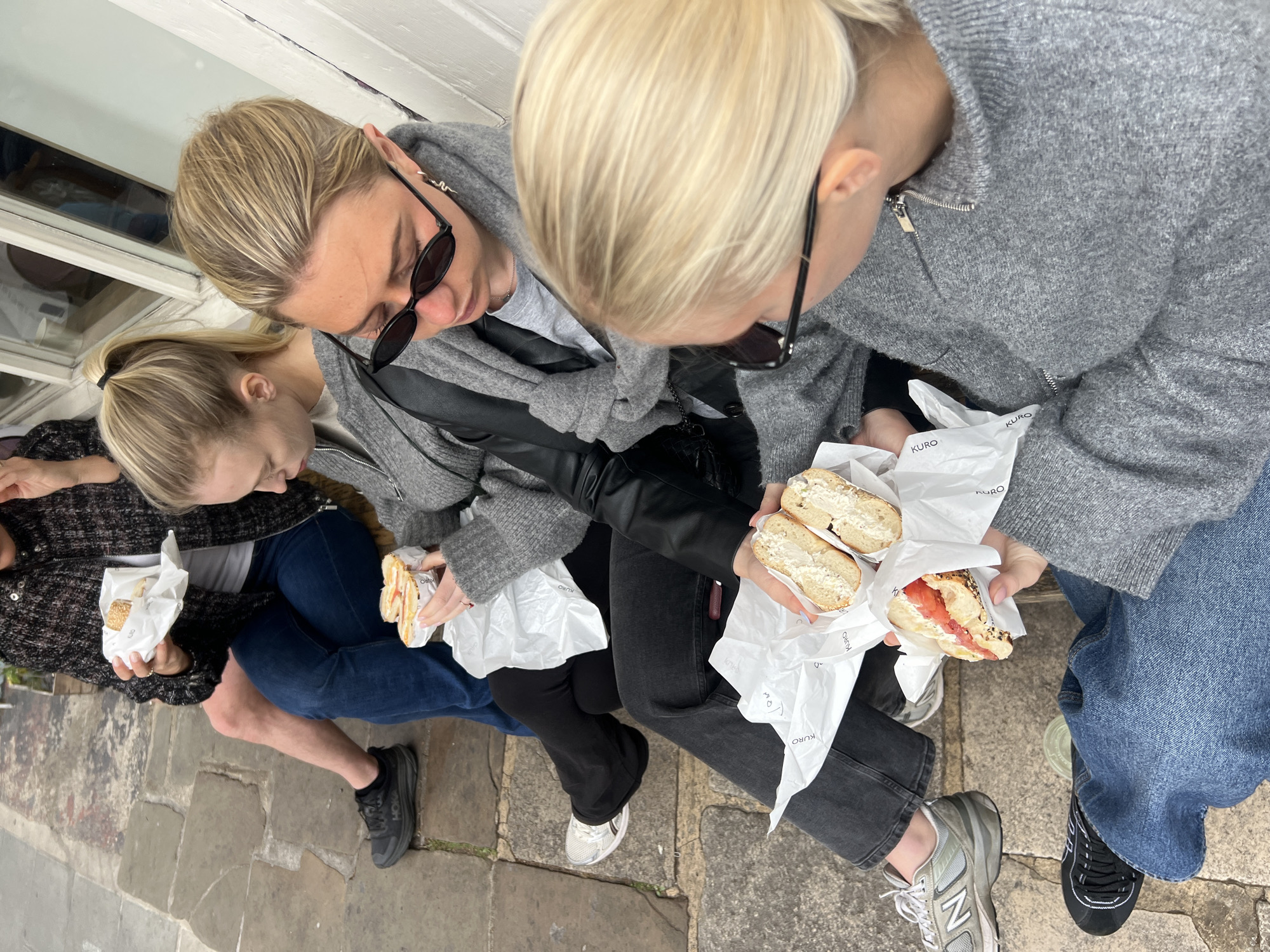 Three young women are sitting outdoors, eating bagels. Two of the women have blond hair pulled back, and the third's hair is the same color but loose. All three are looking down at their food. The woman in the middle is wearing sunglasses and a grey hoodie over a black leather jacket. The woman on the right is wearing a grey hoodie and jeans. The woman on the left is wearing a dark, patterned jacket and jeans. They are all holding bagels in white paper wrappers with the word "KURO" printed on them. The bagels appear to be filled with cream cheese and/or salmon. The women are sitting on a stone ledge or step in front of a white building with visible window frames. The setting is urban, most likely in a European city, given the architectural style and street surface. Based on the branding on the wrapper, this could be the Kuro Eatery located in London, United Kingdom.