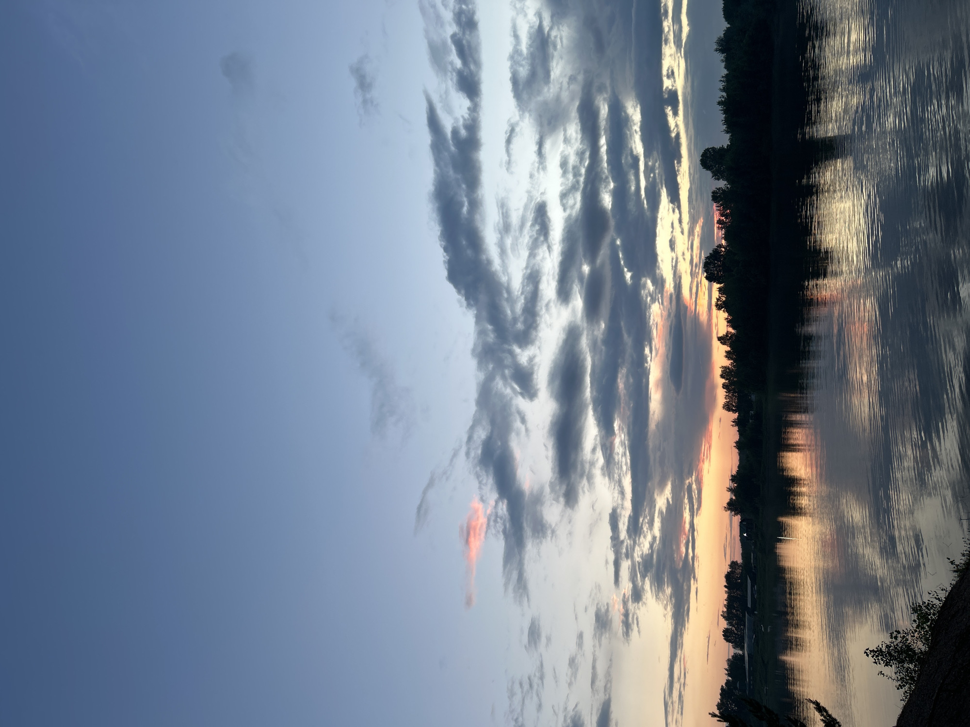 The image depicts a serene natural landscape with a wide river in the foreground, reflecting the dusky sky. The sky is a gradient of soft blues and grays, transitioning to warm oranges and pinks near the horizon, indicating either sunrise or sunset. Scattered clouds are present, some catching the ambient light. Across the river, a dense line of silhouetted trees forms the horizon, with hints of man-made structures, possibly buildings or infrastructure, visible behind them. The river surface is rippled, creating distorted reflections of the sky and the treeline. The overall atmosphere is peaceful and tranquil, suggesting a remote or rural setting. The location is identified as Noviy, Russia. There are no people or animals visible in the scene.