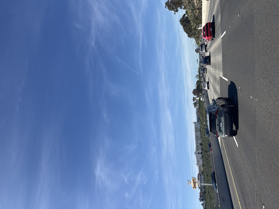 A highway is depicted under a bright blue sky with wispy clouds. Multiple lanes of traffic are visible with various vehicles, including a red car, a dark SUV, and others further down the road. The shoulder of the highway shows some vegetation, a building, and a Denny's restaurant sign. The overall scene conveys a typical highway view on a clear day.
