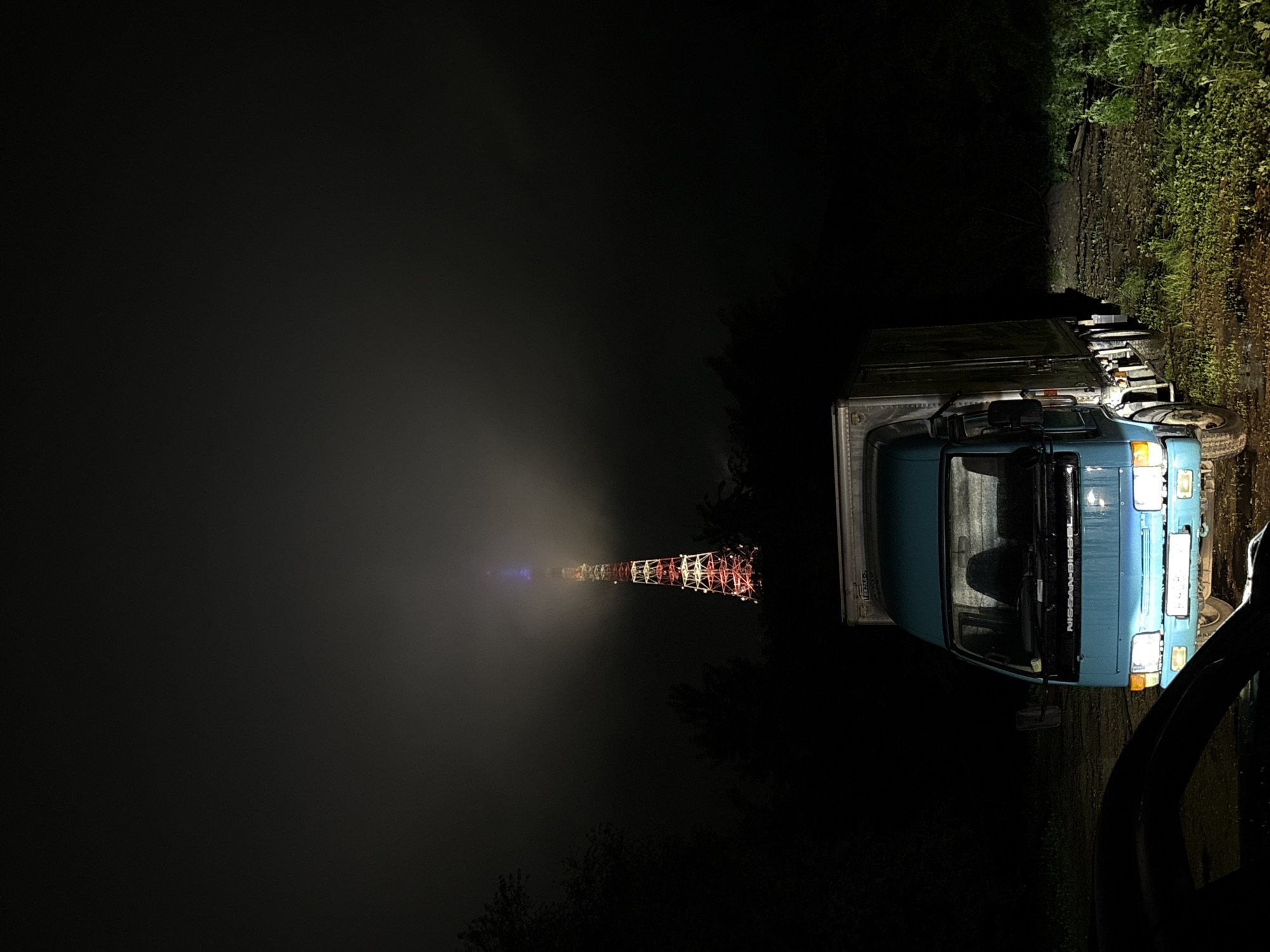The image, captured at night in Birobidzhan, Russia, depicts a dark, foggy outdoor scene with a parked truck and a distant, illuminated tower. The sky is largely obscured by dense mist or fog, which diffuses available light sources.
On the right side of the frame, a blue Nissan Diesel box truck is parked, likely on a muddy or wet dirt track. Its headlights are illuminated, casting a yellowish glow on the immediate foreground. The text "NISSAN DIESEL" is clearly legible in white letters on the truck's windshield visor. The cargo section of the truck is a plain, light-colored box. To the right and behind the truck, lush green vegetation and trees are dimly visible, suggesting a rural or semi-rural environment.
Towards the center-left, in the mid-ground, a tall lattice tower rises through the fog. The tower is brightly illuminated in a striking array of colors, including red, white, and blue. The surrounding fog enhances the tower's lights, creating a glowing halo effect around its structure and an ethereal appearance against the dark backdrop. A distinct beam of blue light emanates upwards from the tower's apex, piercing through the thick mist.
No people are visible in the scene, and the truck appears to be stationary, possibly indicating a stop or parking for the night. The pervasive darkness, combined with the heavy fog and the solitary artificial lights, creates a quiet and somewhat mysterious atmosphere.