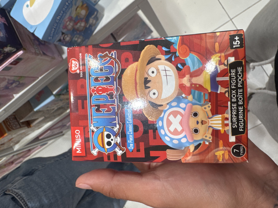 A human hand is holding a red MINISO-branded "One Piece Classic Collection" surprise box figure in a retail store environment. The box prominently features illustrations of the characters Monkey D. Luffy and Tony Tony Chopper. The text "SURPRISE BOX FIGURE," "FIGURINE BOITE PIOCHE," "7 TYPES," and "15+" is visible on the lower portion of the box. "TOEI ANIMATION" is also visible in the upper right. The background consists of white retail shelving displaying various other boxed merchandise, including a blue box with "Sanrio character" text and a cartoon duck design. A section of blue jeans and a white drawstring hoodie are visible at the bottom of the frame, indicating the presence of another individual or the lower body of the person holding the box. The scene takes place within a MINISO store located in León, Mexico.