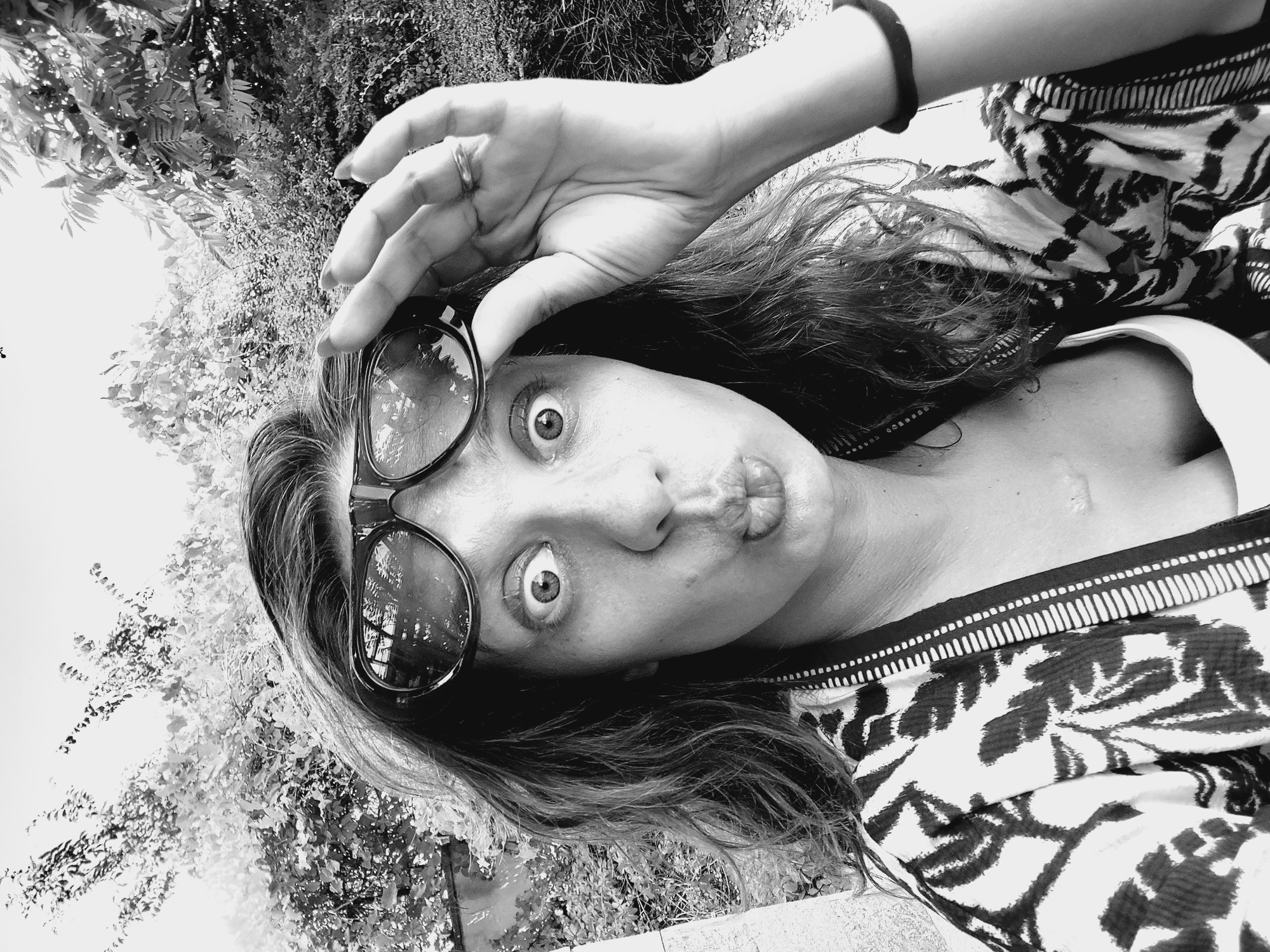 A black and white, close-up, high-angle, selfie-style shot shows a woman's face as she pulls her large-framed sunglasses down her nose, making a fish face with her lips and wide eyes. She has long, wavy, dark hair and is wearing a patterned shirt. Her right hand, adorned with a ring, is raised to her forehead, holding the sunglasses. The background is blurred foliage, suggesting an outdoor setting. The image does not contain any information about the location being the city of Sozopol, Bulgaria.