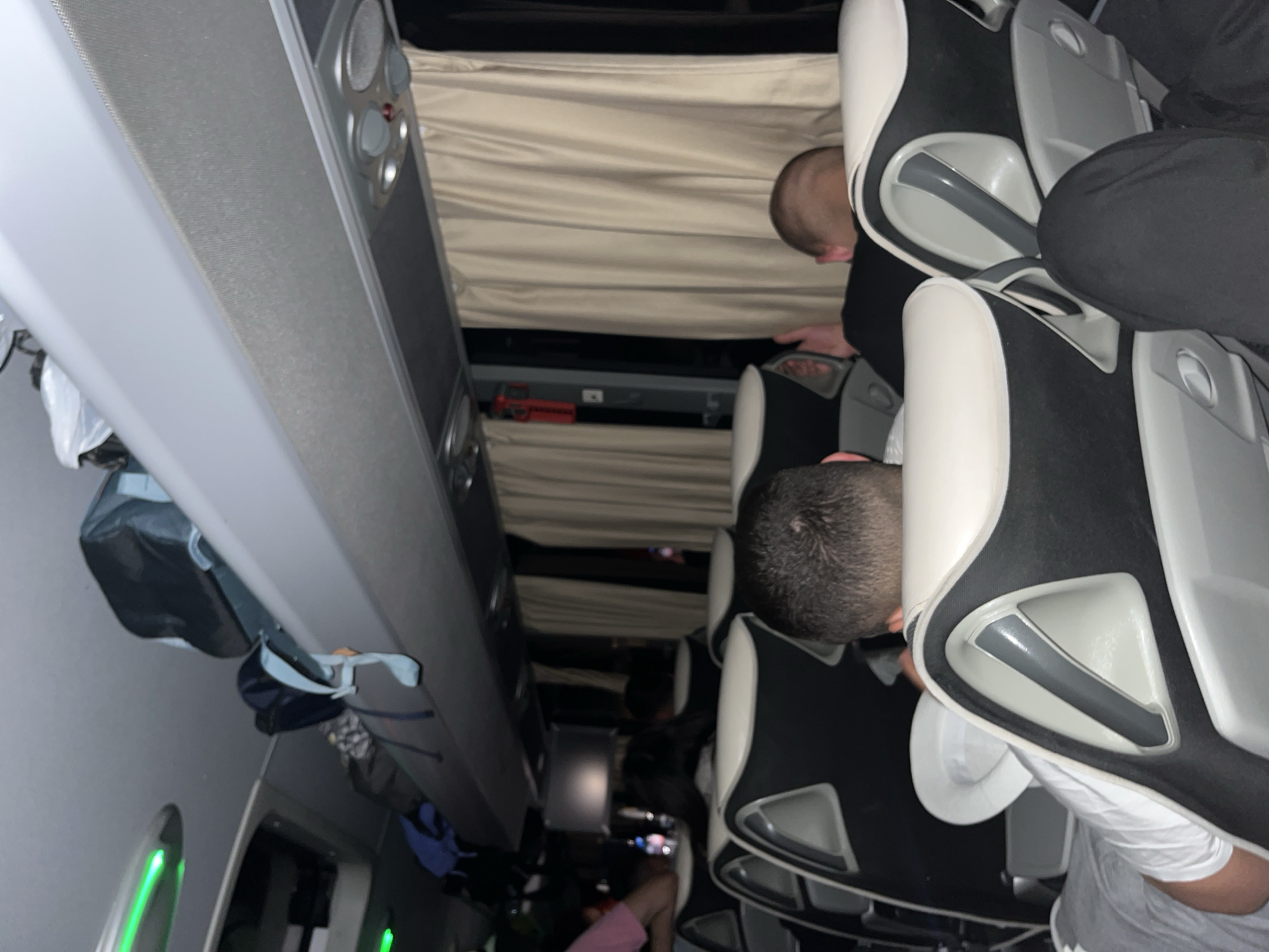 The image depicts the interior of a bus. Passengers are seated in black and white seats, facing forward. Some passengers are visible from behind, including one man reaching under the seat in front of him. Curtains are visible along the side of the bus. Baggage compartments are located above the seats. The location of the bus in Dinkata, Bulgaria, is not verifiable from the image.