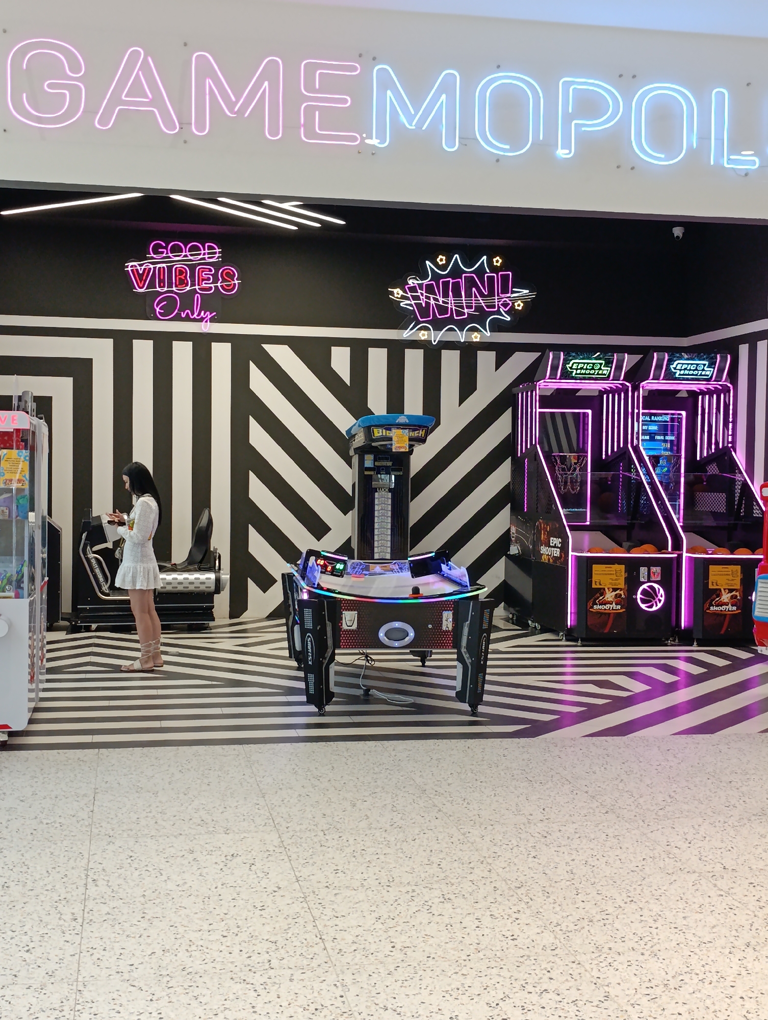 The image shows the interior of an arcade with neon signs and various arcade games. A large neon sign across the top reads "GAMEMOPOL". Below this, two smaller neon signs read "GOOD VIBES Only" and "WIN!". The walls behind the signs have a black and white striped pattern.
In the center of the image, a young woman wearing a white dress and sandals is standing and looking at her phone. She is positioned in front of a large, black arcade game with a seat. To the right of this game are two "EPIC SHOOTER" basketball arcade games with purple neon lighting. To the left of the woman, a claw machine arcade game is visible. The floor is a patterned black and white design, transitioning to a speckled grey and white tiled floor in the foreground. The overall impression is a modern and brightly lit entertainment venue. The image was taken in Chișinău, Moldova.