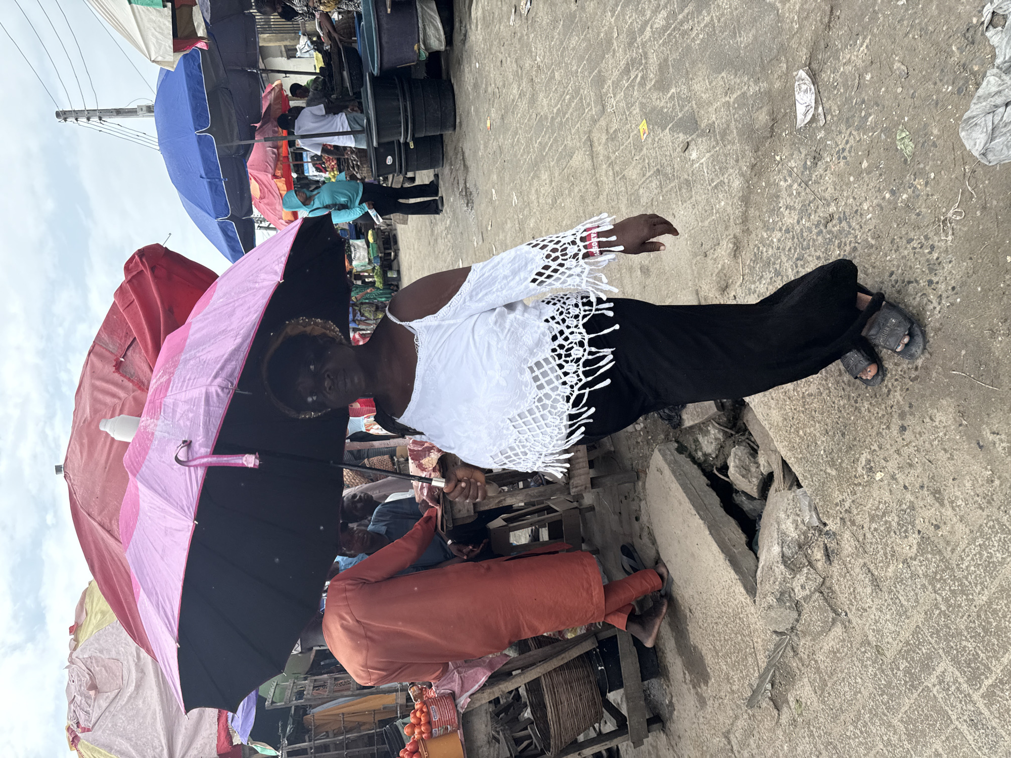 In the city of Ado, Nigeria, a woman in a white off-the-shoulder top and black pants, holding a pink and black umbrella, walks past a market stall. She is wearing sandals and a red band on her left wrist. In the background, other market stalls are visible with various colored umbrellas overhead. A man in an orange robe stands near a stall with tomatoes. The ground is paved with rough concrete and shows signs of disrepair with a large hole in the surface. The scene appears to be a busy outdoor market with people and goods under numerous umbrellas.