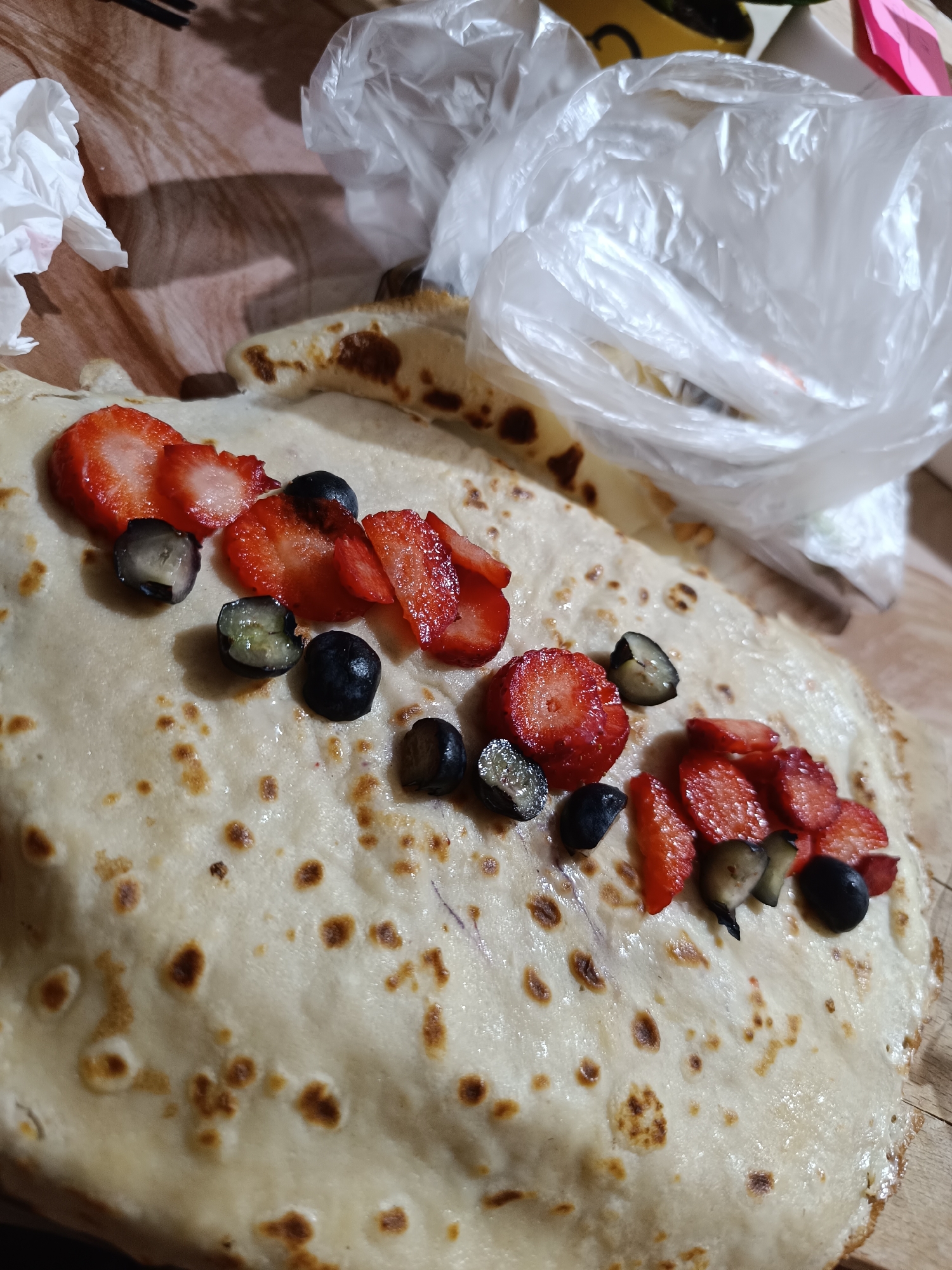 A crepe is displayed on a wooden surface. The crepe is topped with sliced strawberries and whole blueberries arranged in a line. A plastic bag is visible in the upper right corner of the image, and a crumpled napkin is on the left side. The location is Pleven, Bulgaria.