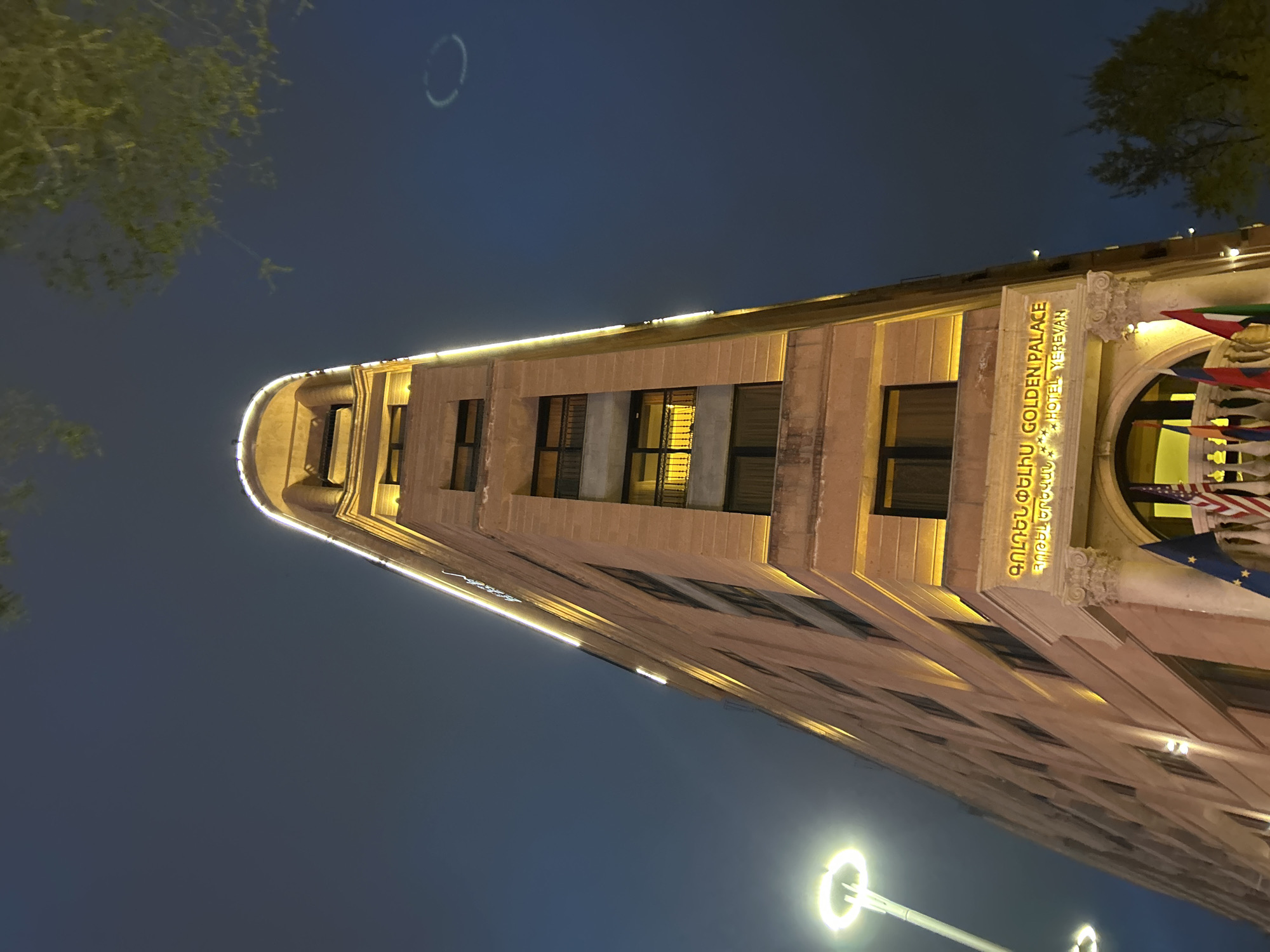The image captures an urban scene at night, dominated by a tall, illuminated building. The building, a hotel, stands prominently with warm, yellowish lights outlining its architectural features and illuminating its windows. Its design incorporates classical or Art Deco elements, featuring decorative cornices and window frames.
Clearly visible on the facade is a large sign that reads "GOLDEN PALACE" in English and "ԳՈԼԴԵՆ ՓԱԼԱՍ" in Armenian script. Below the English name, the text "HOTEL YEREVAN" further specifies the establishment and its location, confirming it to be the Golden Palace Hotel in Yerevan, Armenia. On the right side of the building, several flags are displayed, though their specific designs are not fully discernible.
The sky is a dark blue-black, indicative of nighttime. On the left side of the frame, the dark silhouettes of tree branches with some green foliage are visible, partially obscuring the upper portion of the sky. In the lower left, a modern street light with a distinct circular light fixture emits a bright glow. A faint, circular light or reflection is also noticeable in the dark sky above the building, possibly a lens flare or an unusual atmospheric effect. The overall atmosphere is one of a well-lit, stately building against a dark night sky, typical of a city evening.