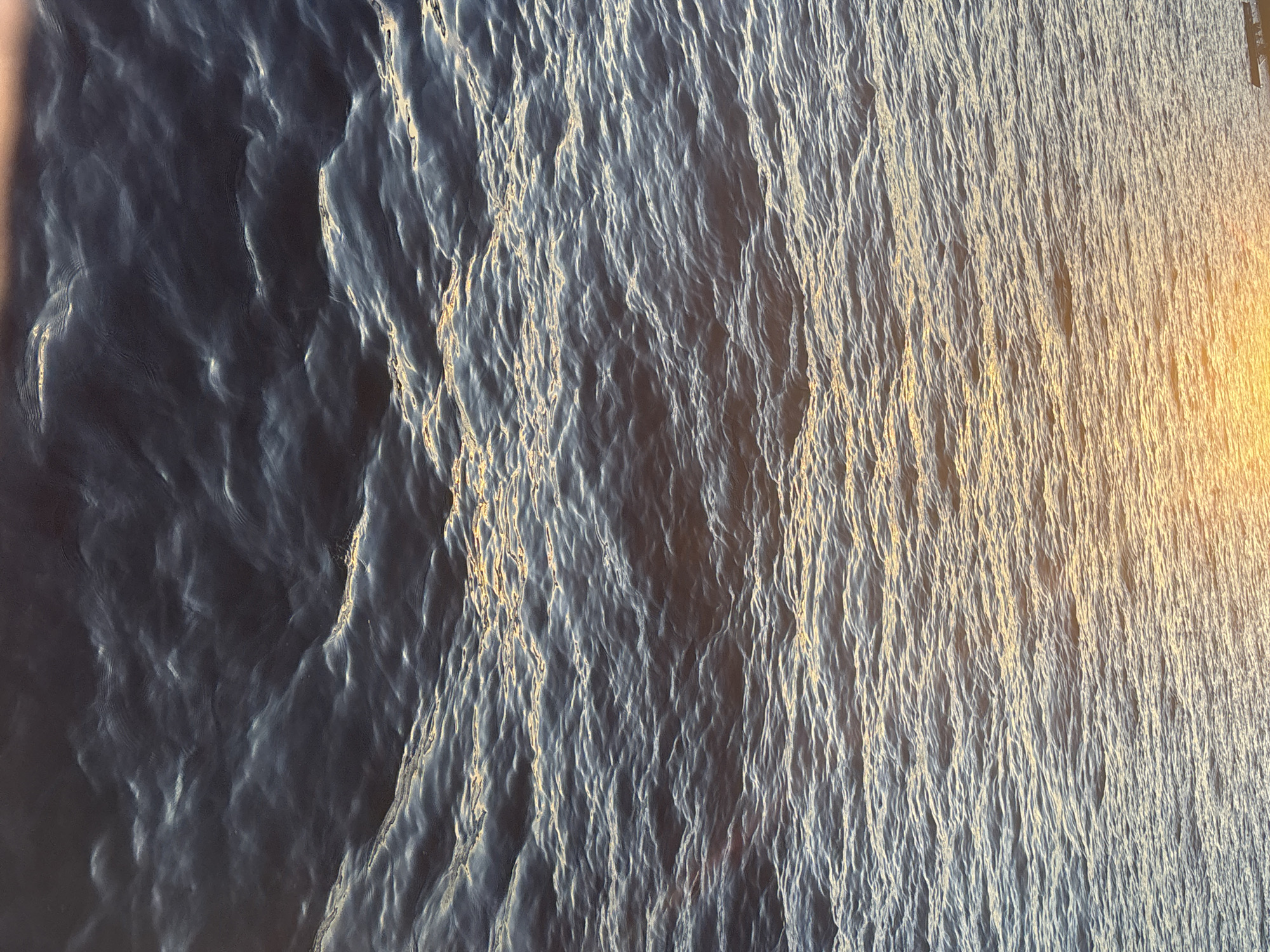 The image shows a close-up view of dark, rippling water, likely a body of water in Saint Petersburg, Russia. The surface of the water is textured with small waves and catches the golden light of either sunrise or sunset, creating streaks of yellow and orange reflections. In the upper left corner, a small portion of a dark object, possibly a boat, is visible at the edge of the water. The overall impression is one of tranquility and natural beauty, characteristic of the numerous rivers, canals, and the Gulf of Finland that are integral to Saint Petersburg's landscape. The water appears to be deep, given its dark color, and the lighting suggests it is either early morning or late evening. There are no people or animals clearly visible in this image.