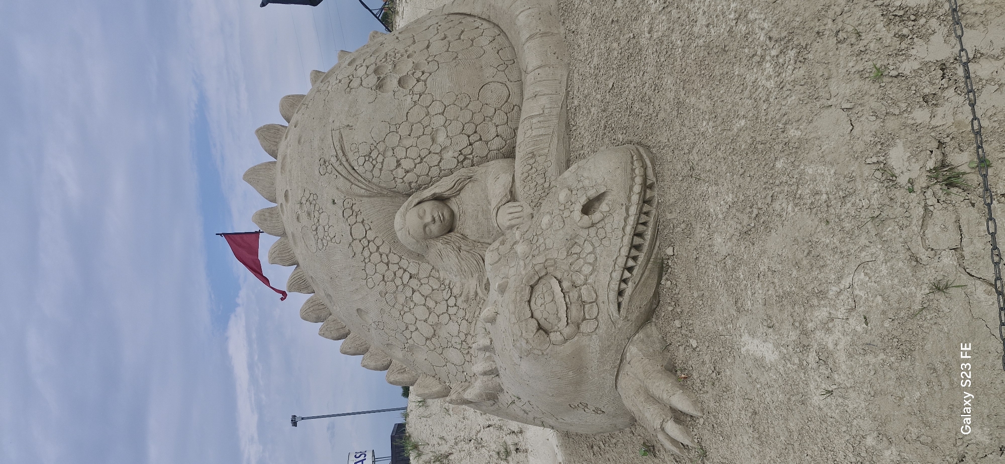 The image depicts a large sand sculpture on a sandy ground under a cloudy sky. The sculpture is of a dragon curled up, with a young girl nestled within its coils. The dragon is rendered with intricate scales and sharp teeth, its eyes closed. The girl appears to be sleeping peacefully, her head resting against the dragon. A red flag is planted atop the dragon's back. In the background, there is a utility pole and a sign, possibly indicating a beach or recreational area. The overall scene suggests a whimsical and fantastical creation on a beach or similar sandy environment. This sand sculpture was created on the bank of the Lielupe river in Jelgava, Latvia, during the International Sand Sculpture Festival.