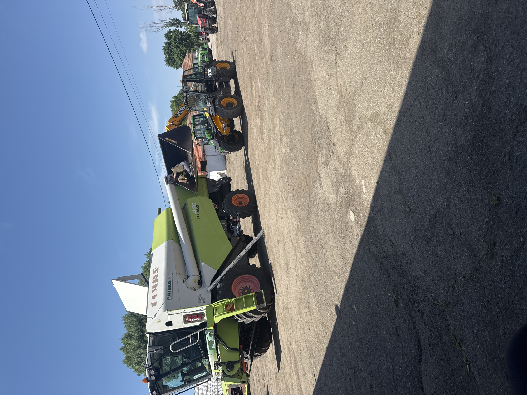 The image depicts a collection of agricultural vehicles on a paved surface. The foreground is dominated by a green and white Claas combine harvester with the model "440 Tucano" visible. Adjacent to the combine, there is a yellow JCB telescopic handler with its boom elevated. Further back, indistinct due to distance, are several other tractors and agricultural vehicles. The location is an outdoor lot on a sunny day. There are overhead power lines visible above the equipment and some greenery can be seen in the background. There are no visible people actively engaged in any activity. The scene has a shadow cast across the pavement indicating a sunny day. There are no features in the photo which allow to specify that this photo was taken in Sarnevo, Bulgaria.