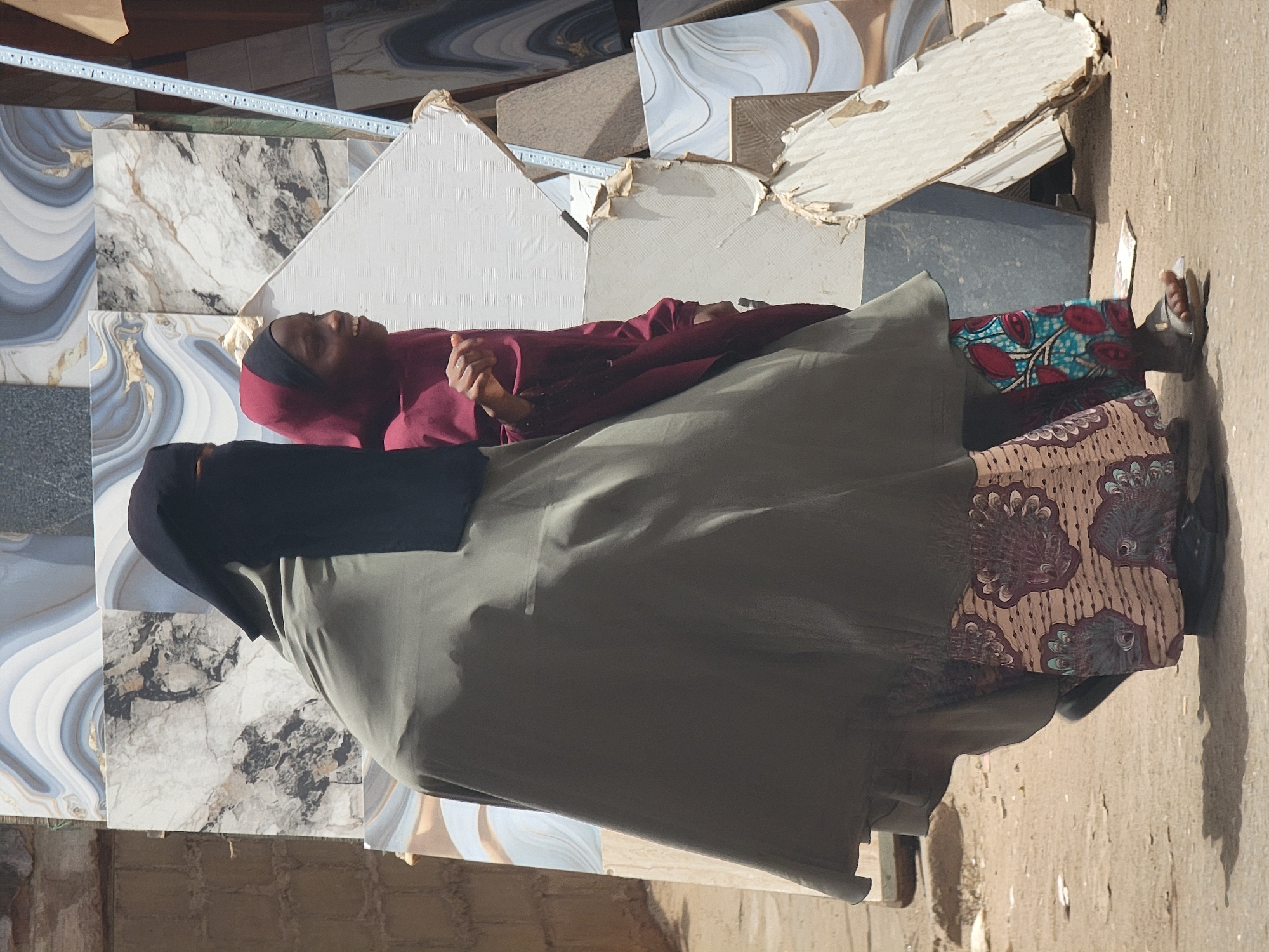 An outdoor scene in Maiburiji, Nigeria, depicts two individuals among construction materials. One figure, positioned towards the left, wears a black hijab and a green gown. Adjacent to them is another person with a red hijab and a maroon dress, adorned with a patterned blue and brown fabric. The second person has a sandal-clad foot visible.
The environment is characterized by an assortment of tiles, including white and grey marble variants, as well as brown-colored tiles. These materials are scattered around the individuals. A partial view of brick construction is seen towards the bottom.