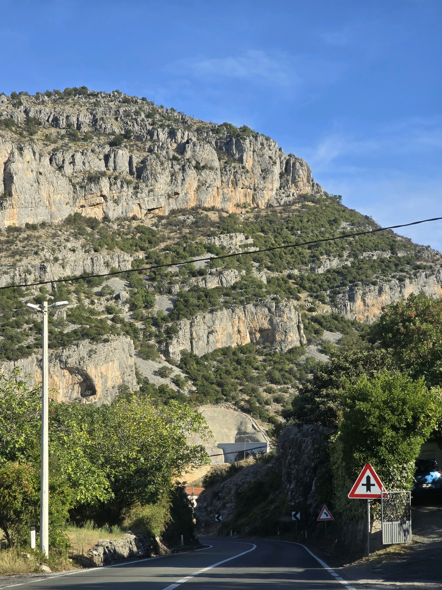 A road curves through a rocky, mountainous landscape under a clear blue sky. The mountain's sheer rock face is textured with layers of grey and tan stone, interspersed with patches of green vegetation. A power line stretches across the mid-section of the frame, strung between poles. A street lamp stands on the left side of the road, and to the right, a white and red triangular road sign indicates a crossroads. Further down the road, another black and white sign shows a sharp bend to the left. Dense green trees and bushes line the sides of the road, obscuring parts of the landscape. In the background, a concrete structure, possibly a tunnel entrance, is visible on the mountainside. A dark-colored vehicle is parked in a driveway on the right side of the image, behind a metal gate. The scene appears to be located on a road leading into or out of a mountainous region near Podgorica, Montenegro.