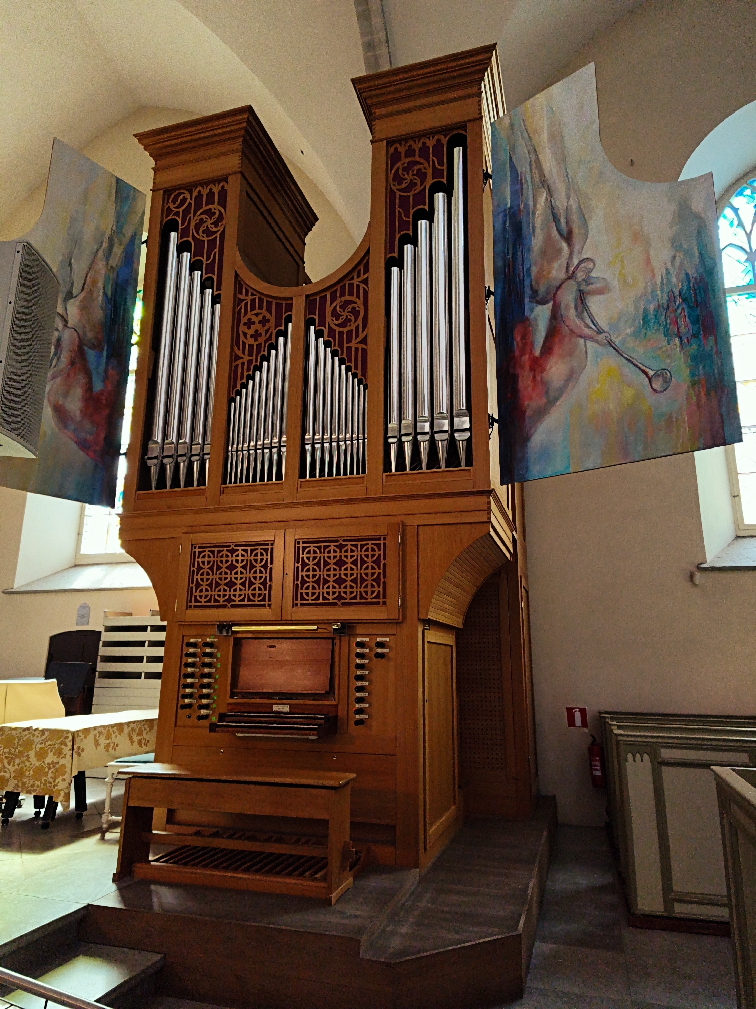 The image shows a large wooden pipe organ in what appears to be a church or concert hall. Two large, abstract paintings are hanging on either side of the organ, featuring depictions of winged figures and musical instruments. The organ itself has numerous pipes of varying sizes, a wooden console with keyboards and stop knobs, and an ornate wooden bench in front of it. In the background, there are tables covered with white and patterned cloths, a window with stained glass, and a fire extinguisher mounted on the wall. The overall setting suggests a place of worship or cultural performance in Tallinn, Estonia, given the context provided.
