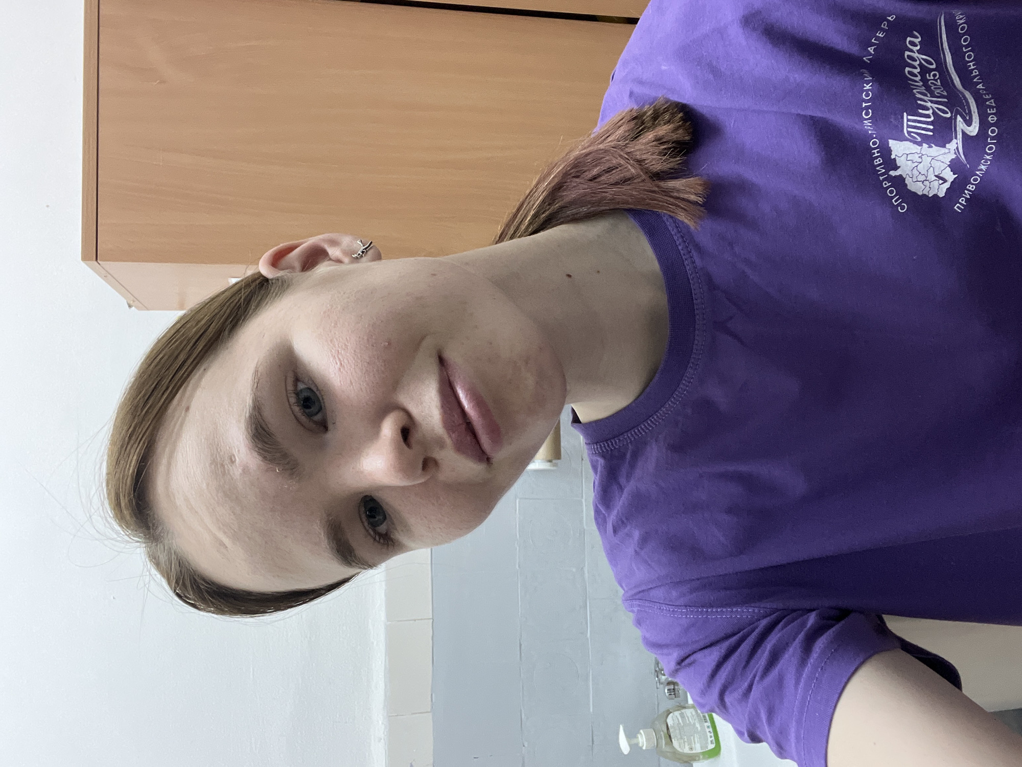A fair-skinned person with blue eyes and light brown hair pulled back into a ponytail is indoors, facing the camera. They are wearing a purple t-shirt with Russian text and a logo. Behind the person is a light-colored wooden cabinet and a white tiled wall. A soap dispenser is visible in the lower portion of the frame. There is no indication that the location is Berezniki, Russia.