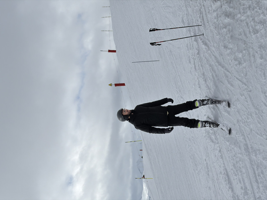 A male individual stands on a snow-covered ski slope in Courchevel, France. He is dressed in a dark ski jacket, dark ski pants, a dark helmet, glasses, and dark gloves, with ski boots fastened to skis. His body is oriented slightly to the left, while his head is turned to his right. Two black and red ski poles are vertically planted in the snow to his right. The background consists of a snowy incline marked by yellow and red ski boundary poles. Distant snow-covered mountains are visible on the left horizon beneath a heavily overcast sky.