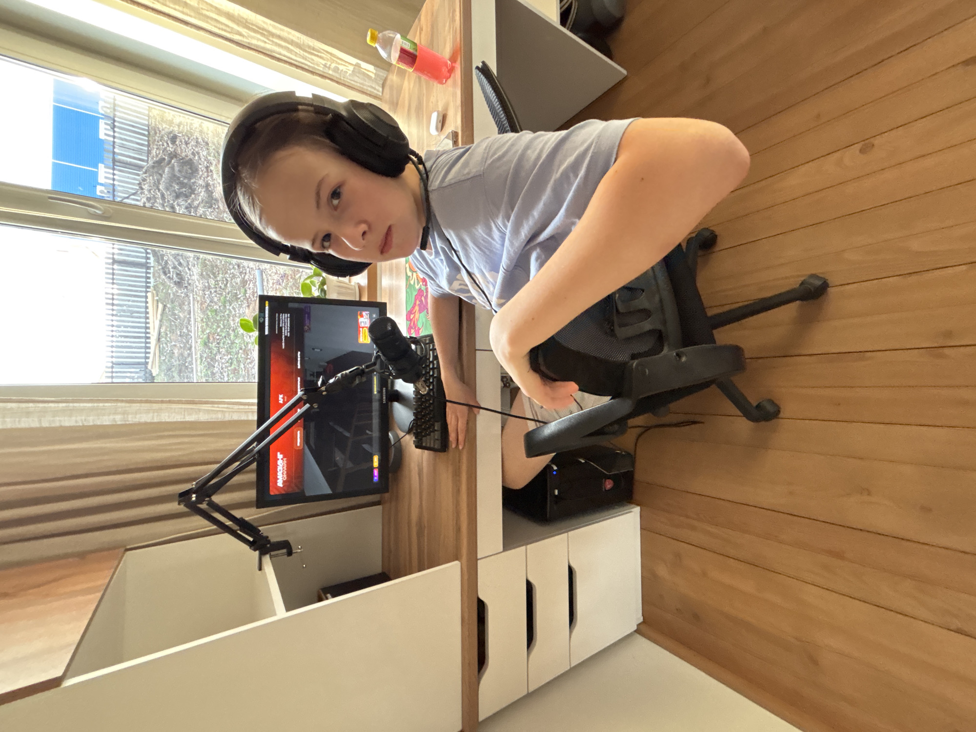 An overhead, tilted photograph captures a young boy seated at a wooden desk in what appears to be a home office or bedroom. He is wearing black over-ear headphones and a light blue t-shirt, looking directly at the camera with a neutral expression.
The desk is equipped with a gaming or streaming setup, including a computer monitor displaying what looks like a video game or an application interface. Partially visible text on the screen includes "SAMSUNG" and "APK", along with fragmented words like "AMAZON" or "AMKSINT". A black microphone on an articulating boom arm is clamped to the desk, positioned near a keyboard. A black PC tower with red accents sits on a lower shelf of the desk unit. To the boy's right, a clear plastic bottle containing a reddish-pink liquid with a yellow cap is on the desk.
Natural light floods the room from a large window to the left, which shows a view of bare trees and a distant structure with blue panels, suggesting daytime. Light-colored blinds are visible on the window. A small green potted plant is also on the desk near the monitor. The room features a wooden floor and white shelving units integrated into the desk. The boy is seated in a black office chair with a mesh back. The perspective is from above and slightly to the boy's right, creating a dynamic, almost "action-cam" feel due to the tilt.