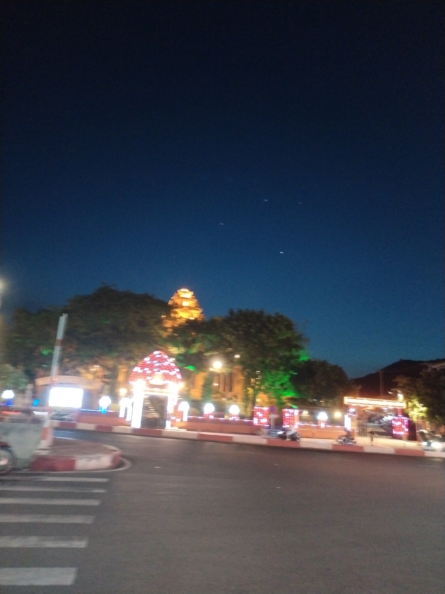 At night in Bac Nha Trang Ward, Vietnam, a road intersection is seen with a lit red-and-white curb and a crosswalk in the foreground. In the background, there is a decorated archway with red and white lights and a brightly lit, multi-tiered building resembling a pagoda. Trees are interspersed with streetlights along a low wall decorated with red and white patterns. Motorbikes are parked along the wall. A mountain is visible on the horizon. The sky is dark blue.