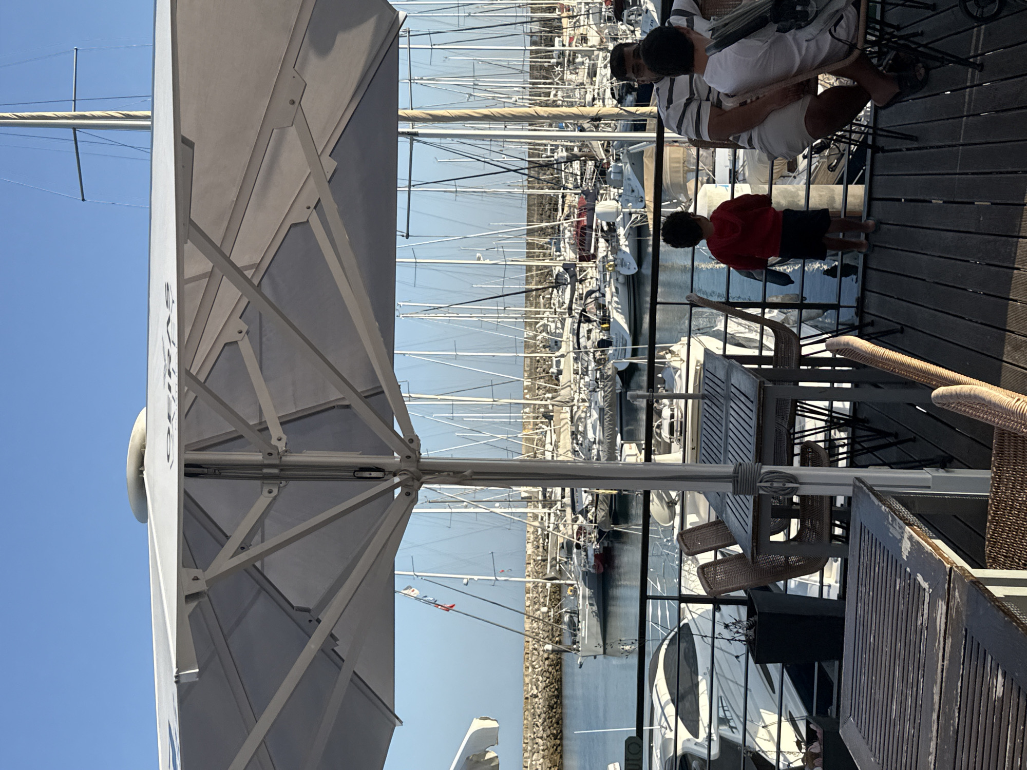 The image depicts an outdoor seating area at a restaurant or cafe, with a large white umbrella providing shade. In the background, a marina filled with numerous sailboats and yachts is visible under a clear blue sky. A stone breakwater separates the marina from the open sea.
In the foreground, there are several wooden tables and chairs, some made of woven material. Three people are seated at one of the tables: an adult male wearing a striped shirt and white shorts, and two younger individuals. One of the younger individuals, a child in a red hooded sweatshirt and dark shorts, stands facing the marina, looking out at the boats. The other younger person, wearing a white t-shirt and dark shorts, is seated.
The seating area is elevated, with a wooden deck floor and a metal railing separating it from the marina. Some potted plants are visible near the railing. The overall scene suggests a relaxed atmosphere in a waterfront location. The presence of the word "OEIRAS" is visible on the umbrella, indicating the location is in Oeiras, Portugal.