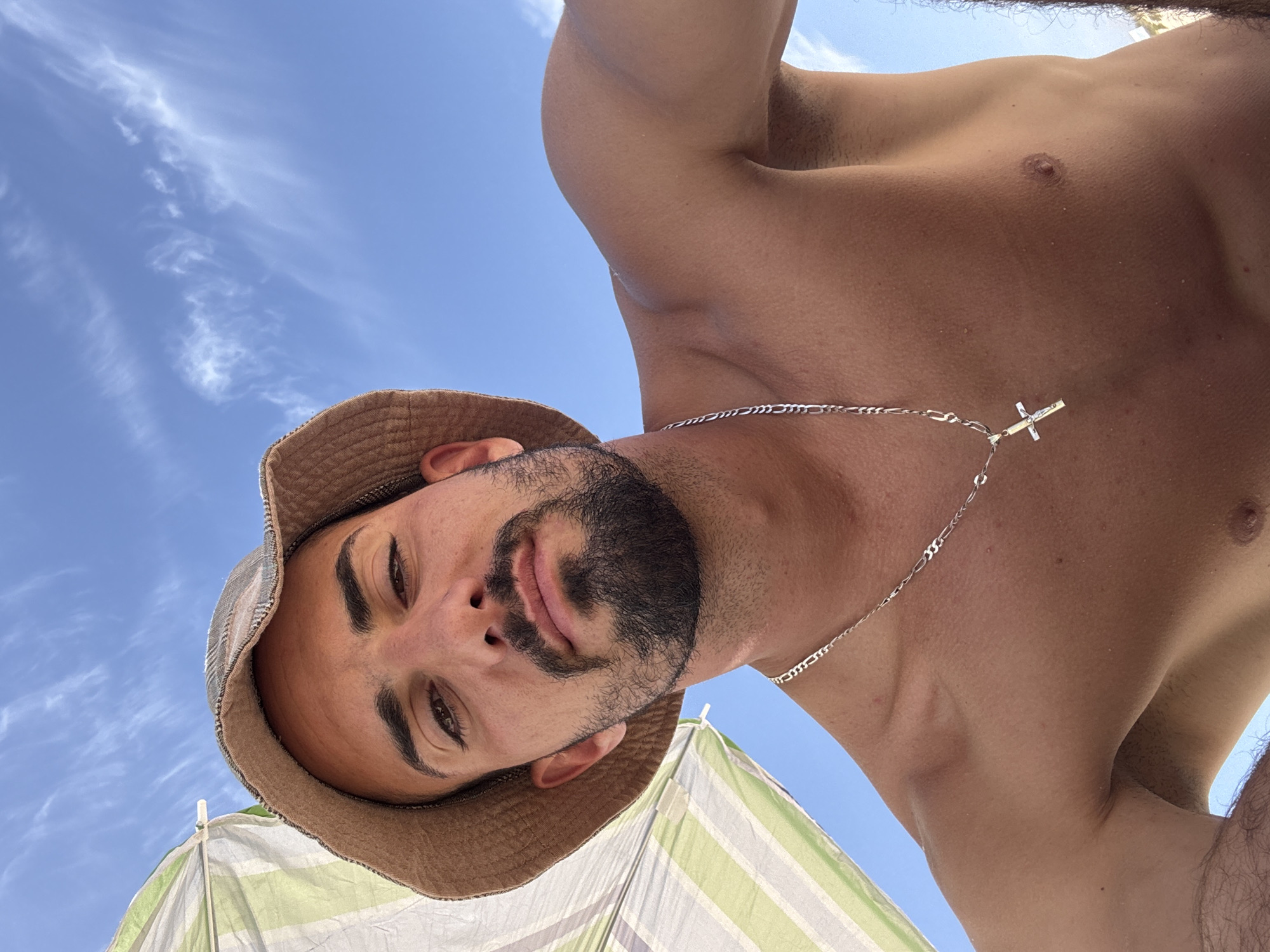 A man is looking down at the camera, wearing a brown bucket hat and a silver chain with a cross pendant. He has a beard and tan skin. The sky is blue with some clouds visible in the background. Part of a green and white striped umbrella is visible in the lower left corner of the image.