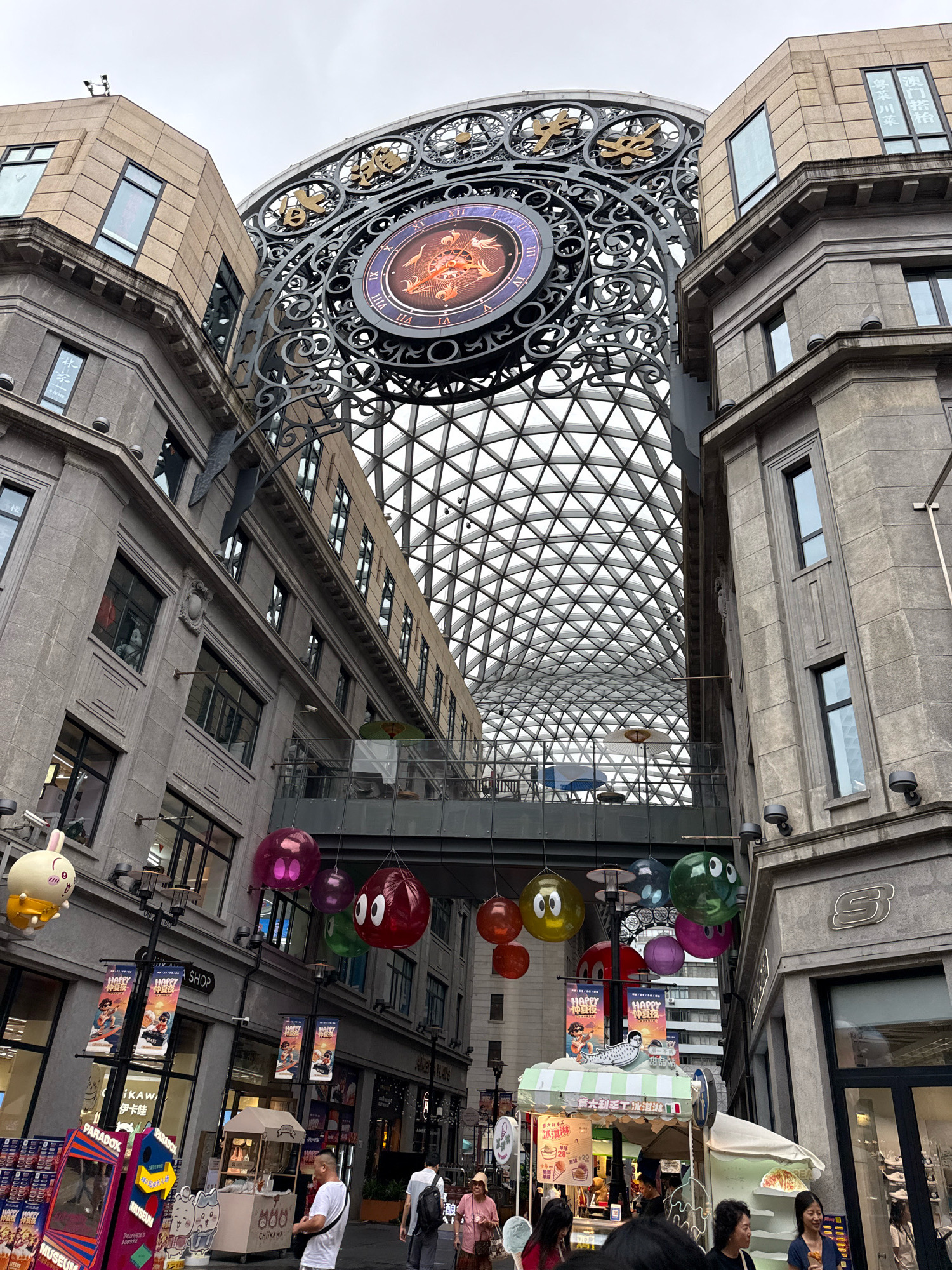 The image depicts the exterior of the Shanghai World Financial Center's "Bonds" area, a shopping street with a distinctive arched glass roof and a large ornamental clock. The architecture is characterized by grey stone buildings with contrasting beige upper sections. On the left, a tan building features multiple windows and a Chinese inscription. The central focus is a large, ornate clock with Roman numerals and an animated dragon design. Below the clock, a modern glass and metal dome structure covers a pedestrian walkway.
The street level is lined with shops and food stalls. Several colorful, inflatable cartoon characters, resembling a rabbit and round faces, are suspended from above. Banners advertising "Happy Night" are visible, along with signage for shops such as "Paradox" and "Skechers." A small food cart selling what appears to be baked goods is in operation.
Several individuals are present on the street. On the left, a person in a white t-shirt and black shorts is near a food cart. Further back, a couple is walking, the woman wearing a pink hat and the man with a backpack. In the foreground, a group of people are gathered, some looking at food stalls. The sky is overcast, suggesting a cloudy day. The overall scene conveys a lively, commercial atmosphere within a modern urban setting.