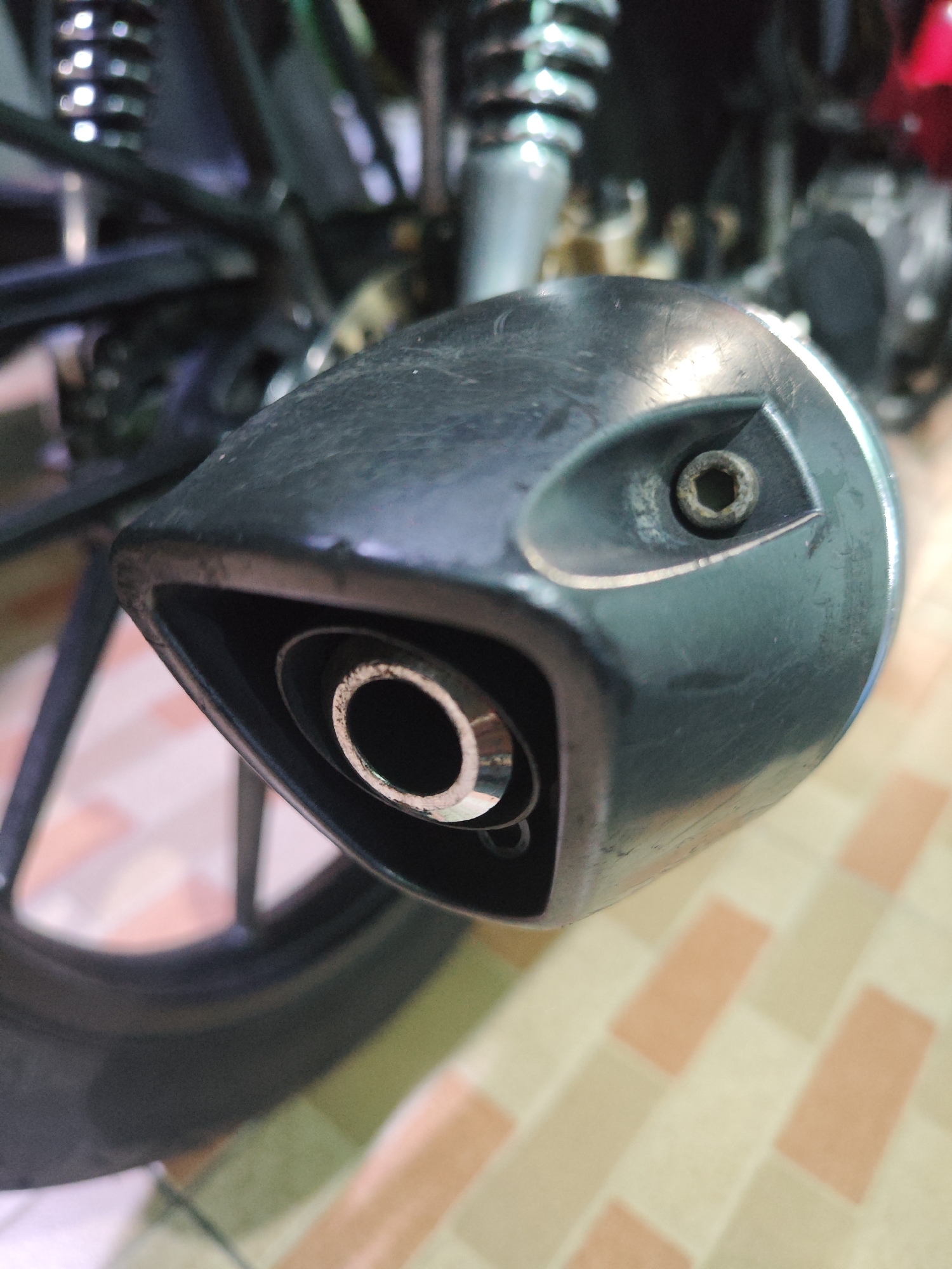 A close-up, low-angle shot captures the exhaust pipe of a motorcycle. The exhaust pipe is black, with a metallic end featuring a circular opening. A silver hexagonal bolt is visible on the side of the exhaust pipe. The motorcycle's black rear tire and suspension are partially visible in the background, along with a red object which might be part of the motorcycle. The flooring beneath the motorcycle appears to be tiled in a pattern of cream and pale orange rectangles. This scene is located in Sroyo, Indonesia.