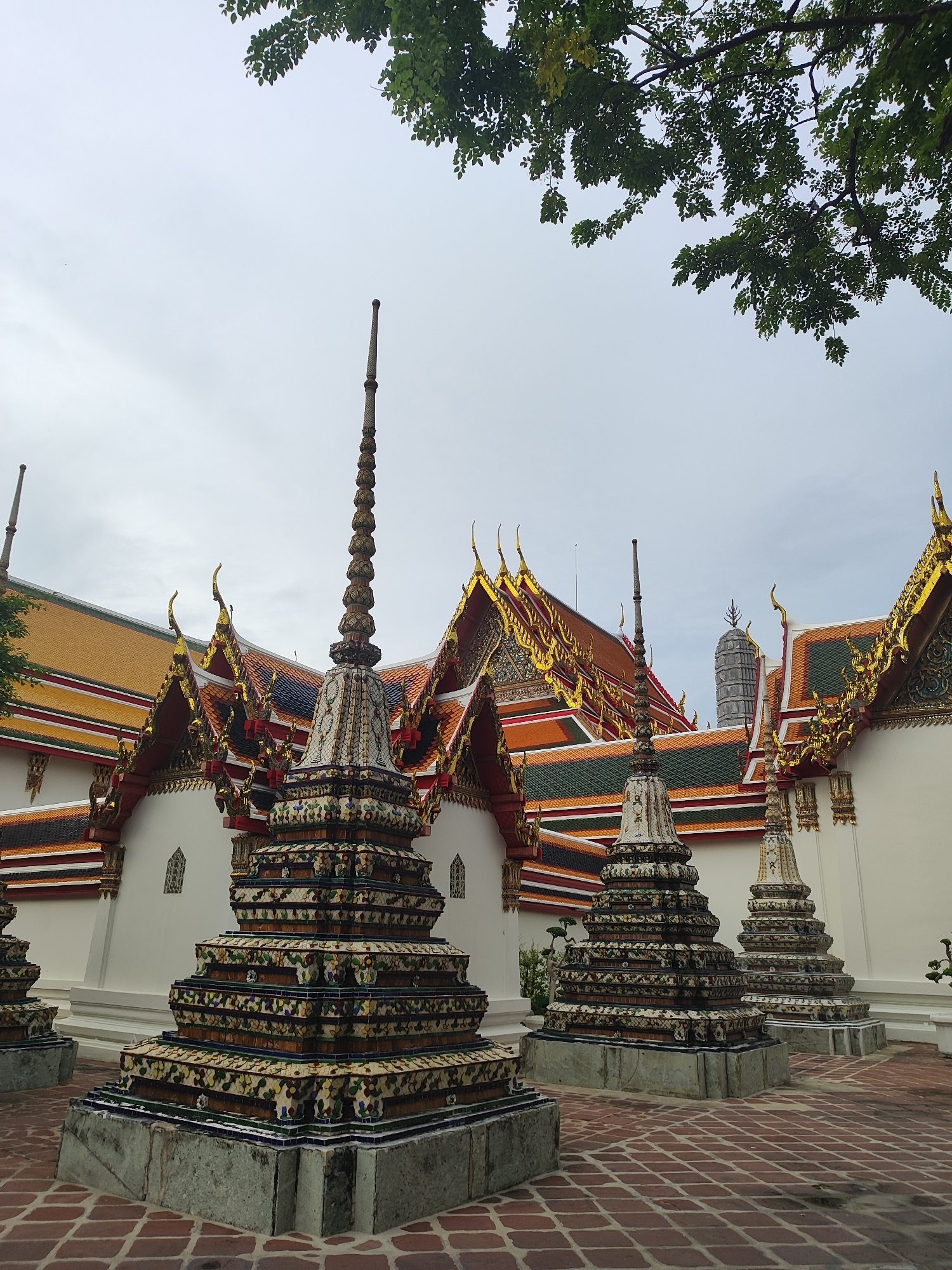This image depicts a view of Wat Pho (Temple of the Reclining Buddha) in Bangkok, Thailand, with its distinctive ornate temple architecture and chedis (stupas). In the foreground, three prominent, multi-tiered chedis are visible, each elaborately decorated with colorful ceramic tiles featuring floral and geometric patterns. The chedis rest on grey stone bases. The ground is paved with red brick laid in a diamond pattern.
Behind the chedis, the multi-tiered roofs of the temple buildings rise, adorned with traditional Thai architectural elements such as gilded finials and vibrant orange, green, and red tiling. Intricate gold detailing runs along the rooflines. The walls of the buildings are white. In the background, a taller, grey, ribbed structure, likely another chedi or tower, can be seen peeking above the main temple roofs.
The sky is overcast and a light grey. The upper portion of the image is partially obscured by the leafy branches of a tree, its green leaves providing a natural frame to the scene. No people are visible in the image. The overall impression is one of serene architectural grandeur in a temple setting.