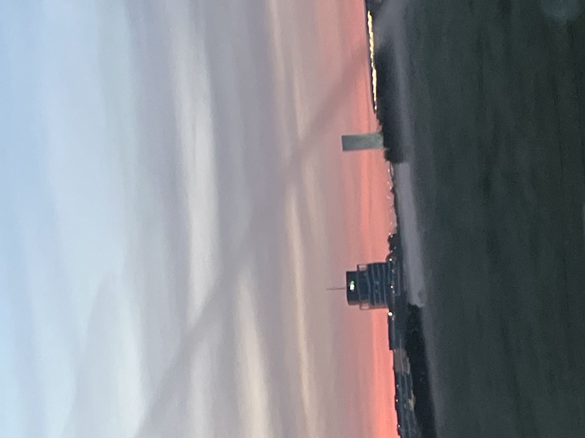 The image captures a hazy sunset over a semi-urban landscape in Leudelange, Luxembourg. In the mid-ground, a distinctive cylindrical building with a rounded top, identified as the "Centre de Recherche Public Henri Tudor," stands tall, illuminated by a green light near its summit. To its right, a slender, rectangular tower is visible, casting a long shadow. A line of lower buildings stretches across the horizon, with some windows reflecting the warm hues of the sunset. The foreground consists of a dark, grassy field, partially obscured by a misty haze that seems to emanate from some unseen source, possibly a sprinkler system. The sky above transitions from a soft blue at the top to a vibrant pink and orange gradient near the horizon, with subtle cloud formations creating a layered effect. The overall atmosphere is serene, with the light pollution from the buildings and the hazy conditions contributing to the diffused light.