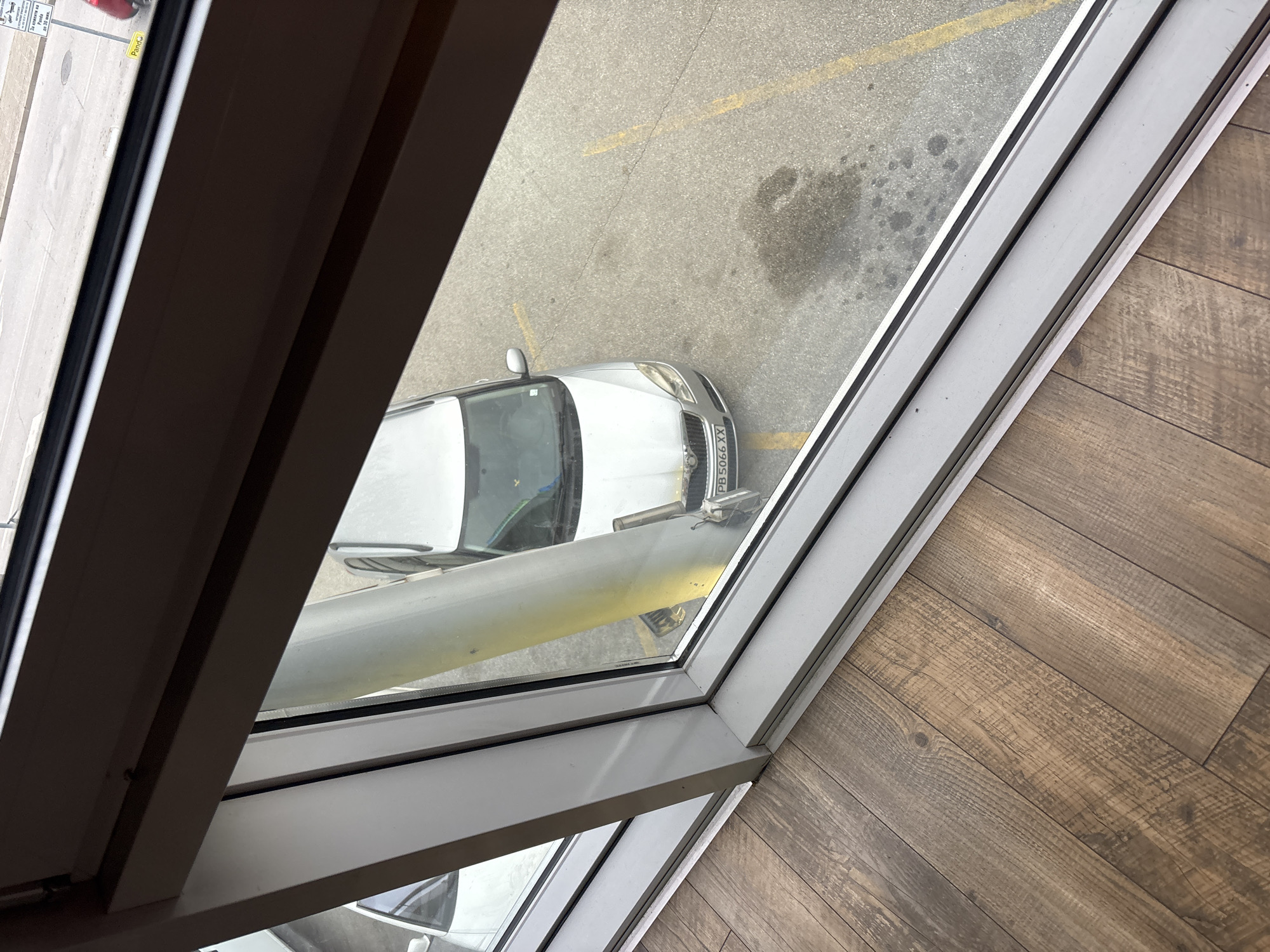 A view from an elevated position, likely inside a building, looks down at a street scene in Plovdiv, Bulgaria. The frame of the window dominates the view. Outside, a silver car is parked partially in view with license plate "PB 5066 XX", suggesting a Bulgarian vehicle registration. The road surface appears to be asphalt with painted yellow lines. Patches of darker stains are visible on the road. The interior space has wood-like flooring. Light reflections on the glass are present.