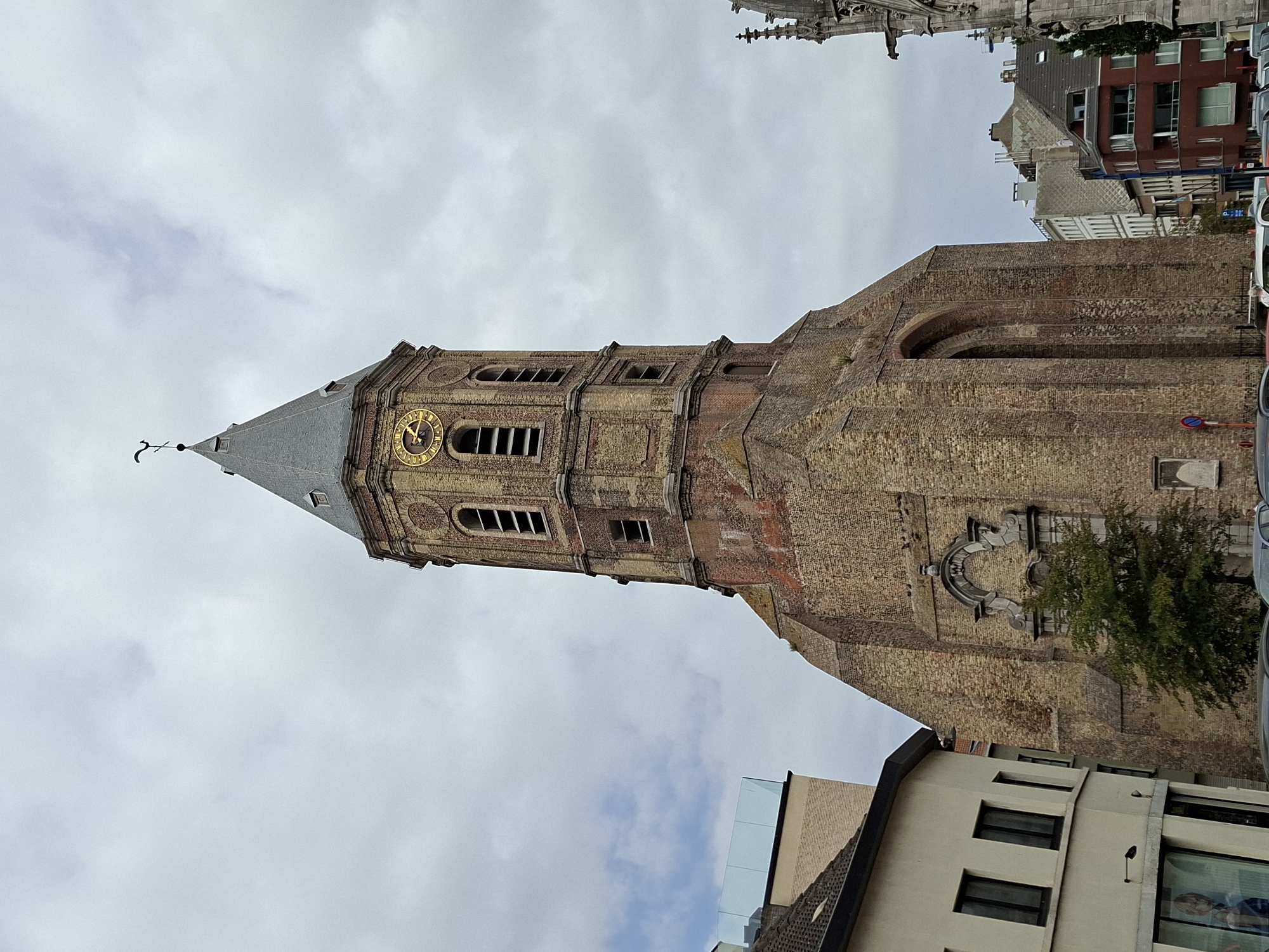 The image captures a view of the Sint-Petrus-en-Pauluskerk (Church of Saint Peter and Paul) in Ostend, Belgium, under a cloudy sky. The church's prominent brick tower, featuring a clock with Roman numerals and a weather vane, dominates the frame. The tower has multiple levels with arched openings for bells. To the left of the church, a modern building with a glass railing is visible, juxtaposed against older brick architecture. In the foreground, cars are parked along the street, and a small green tree grows near the base of the church. The overall scene depicts an urban environment with historical and modern architectural elements.