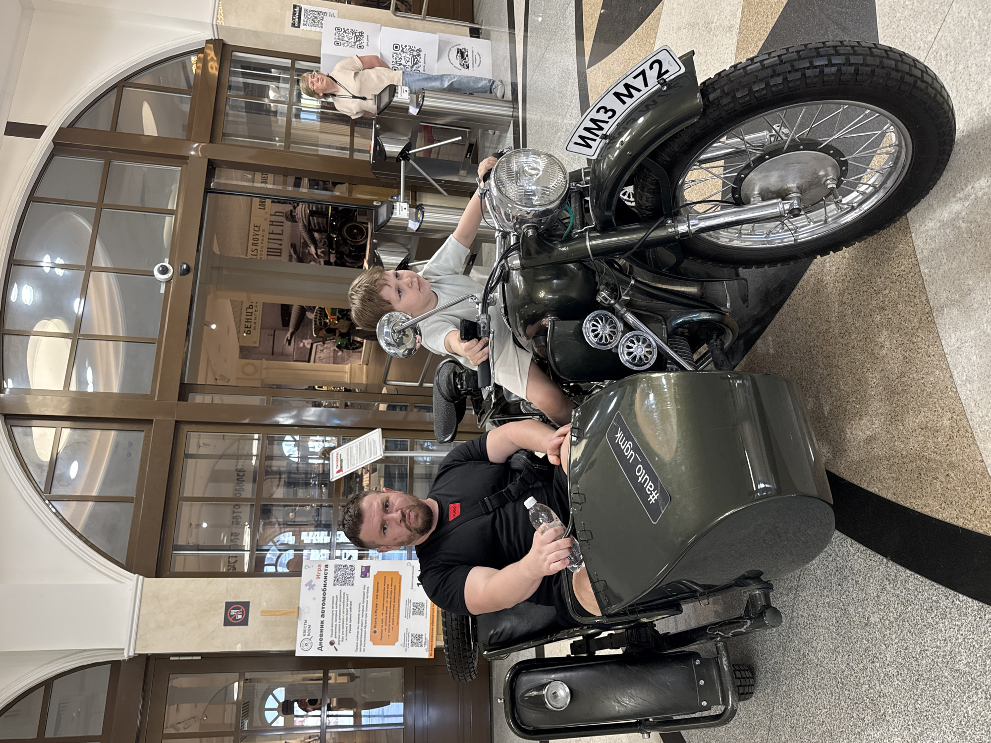 The image captures a scene at the Museum of Automotive Technology in Verkhnyaya Pyshma, Russia. A man is seated in the sidecar of a dark green motorcycle with a sidecar, while a young boy sits on the motorcycle itself. The motorcycle has a license plate that reads "ИМЗ М72". The man is holding a water bottle and wearing a black t-shirt with a cross-body bag, while the boy is in light-colored clothes. Behind them, there's a glass display with text in Russian, including "Бенц" and "Мангнйл", along with another sign about "Дневник автомобилиста" (Driver's Diary). A woman in a beige shirt and jeans is standing in the background. The environment appears to be indoors, with tiled flooring and brown-framed glass walls. The sticker with hashtag #auto_ugmk is on the sidecar.
