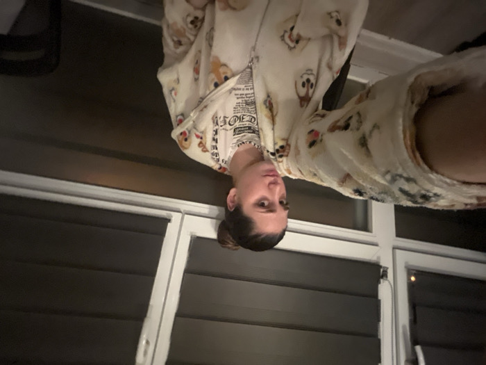 A woman in a light-colored, patterned onesie stands indoors. She also wears a white t-shirt with writing. Her dark hair is pulled back in a bun. Behind her is a wall with horizontal paneling and windows with white frames. The image is rotated so the woman appears upside down.