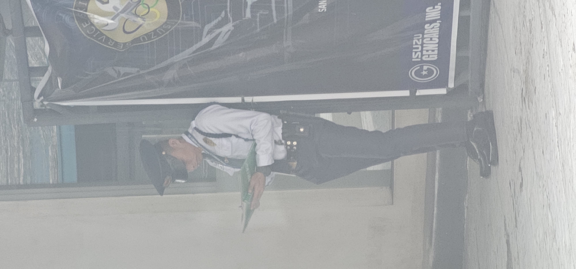 A security guard, wearing a white collared shirt, dark pants, a peaked cap, and a lanyard with an ID badge, stands by a gate and a dark banner. The guard is looking down at a green folder or clipboard they are holding in their hands. The banner has text and logos on it, including "ISUZU GENCARS, INC." and "SAN". The ground appears to be paved. The overall lighting suggests an outdoor environment, possibly during the day. The image quality is low, with a hazy, washed-out appearance.