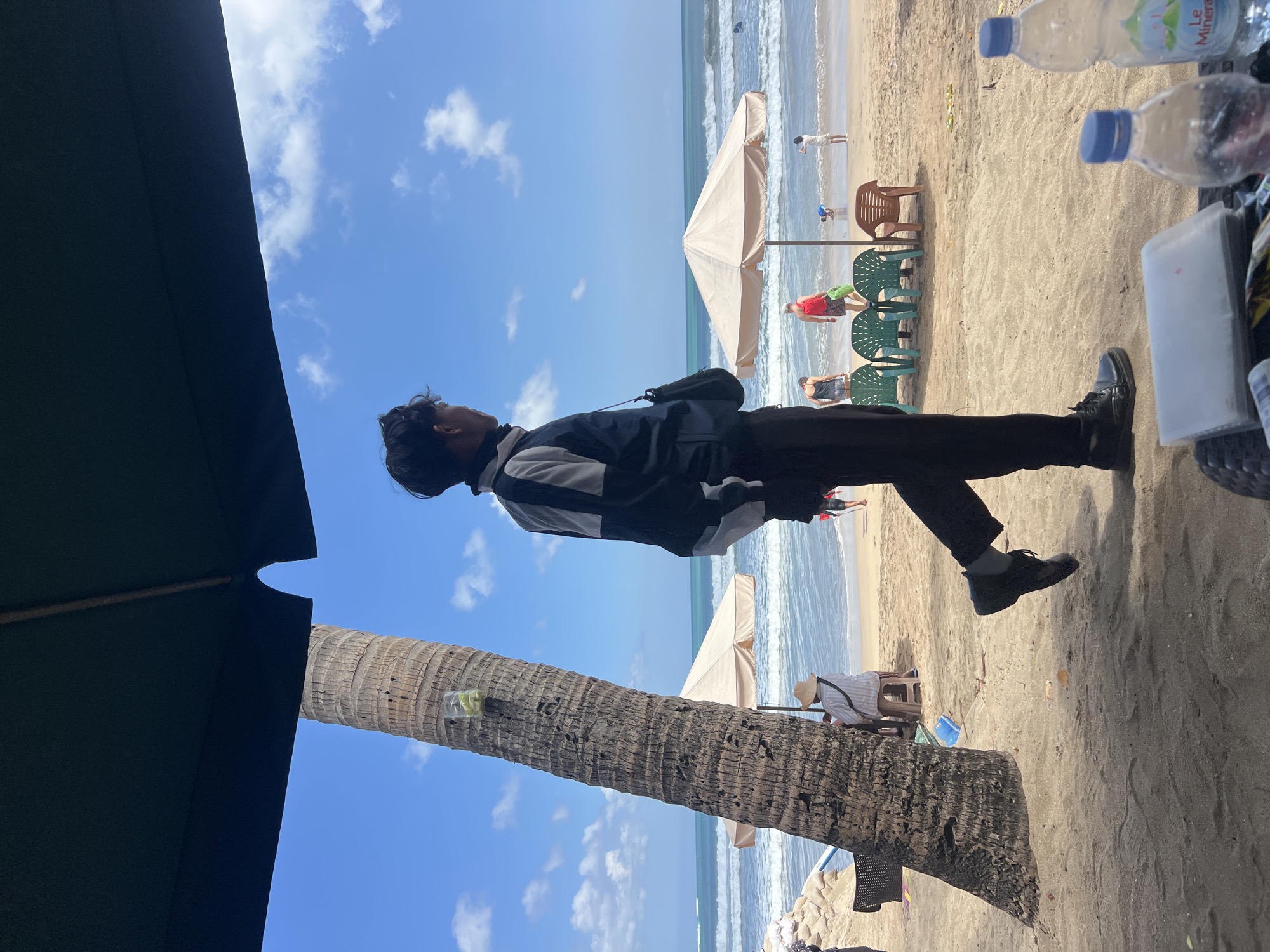 This image depicts a beach scene in Kuta, Indonesia, under a large dark green umbrella. In the foreground, a young man in a black and gray jacket and black pants is walking on the sand, facing right. He has a black bag slung across his shoulder. To his left, a tall, textured palm tree stands prominently. Beyond the palm tree and the man, the sandy beach continues towards the ocean. Several beach umbrellas are set up on the sand, with chairs placed in front of some. In the distance, people are visible on the beach and in the turquoise water, with waves breaking near the shore. The sky is blue with scattered white clouds. In the immediate foreground, out of focus, are bottles of water and other items on a table or surface.