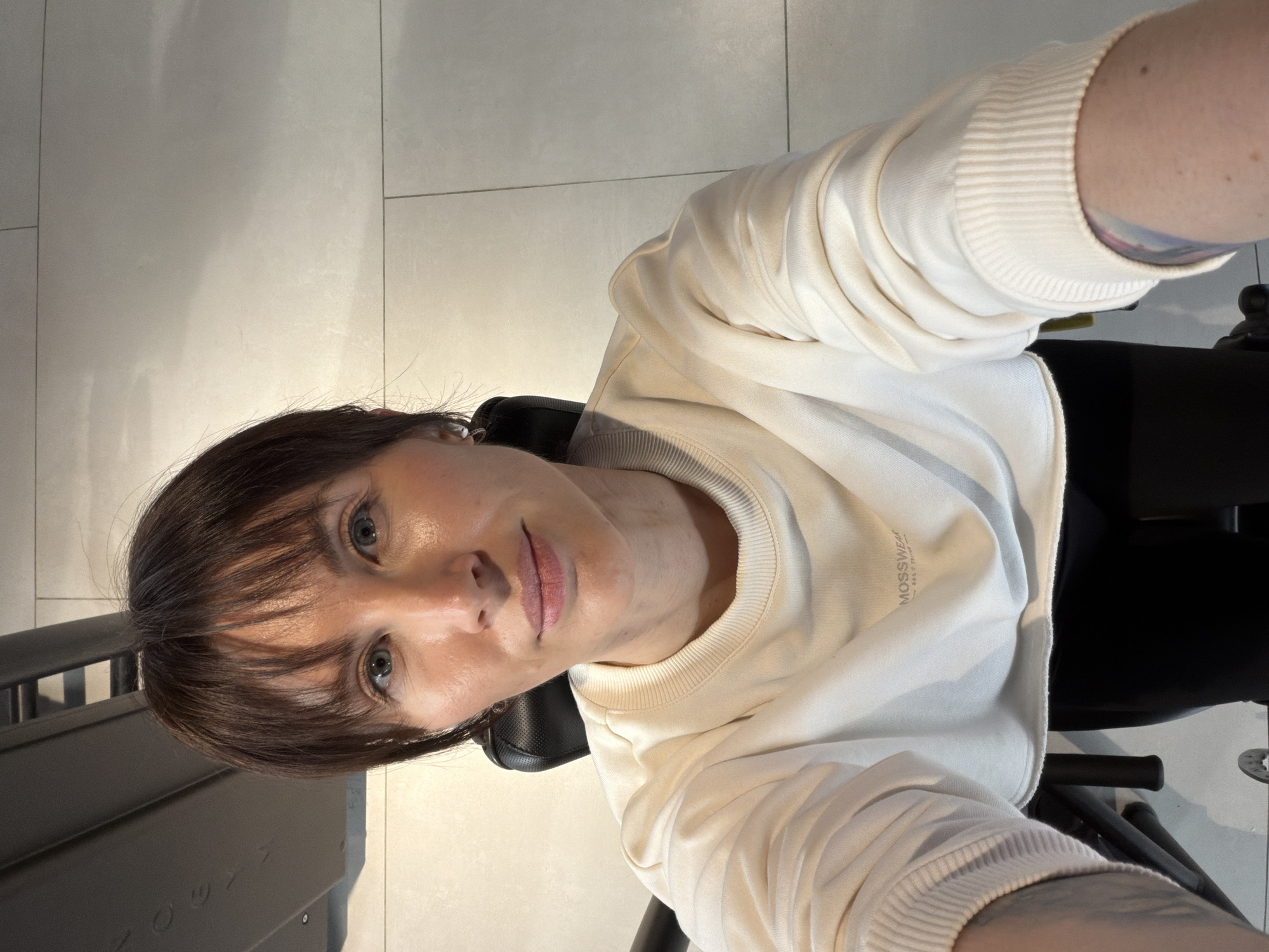 A woman with brown hair and bangs, wearing a cream-colored sweatshirt with the word "MOSSWEAR" printed on it, is seated in a black chair. The woman's left arm, which is tattooed, is partially visible in the upper right corner of the image, while the sweatshirt is partially visible in the bottom-right corner as well. The background consists of light-colored tiled flooring. The image appears to have been taken from a slightly elevated angle. The exact location in Perm, Russia, cannot be determined from the image alone.