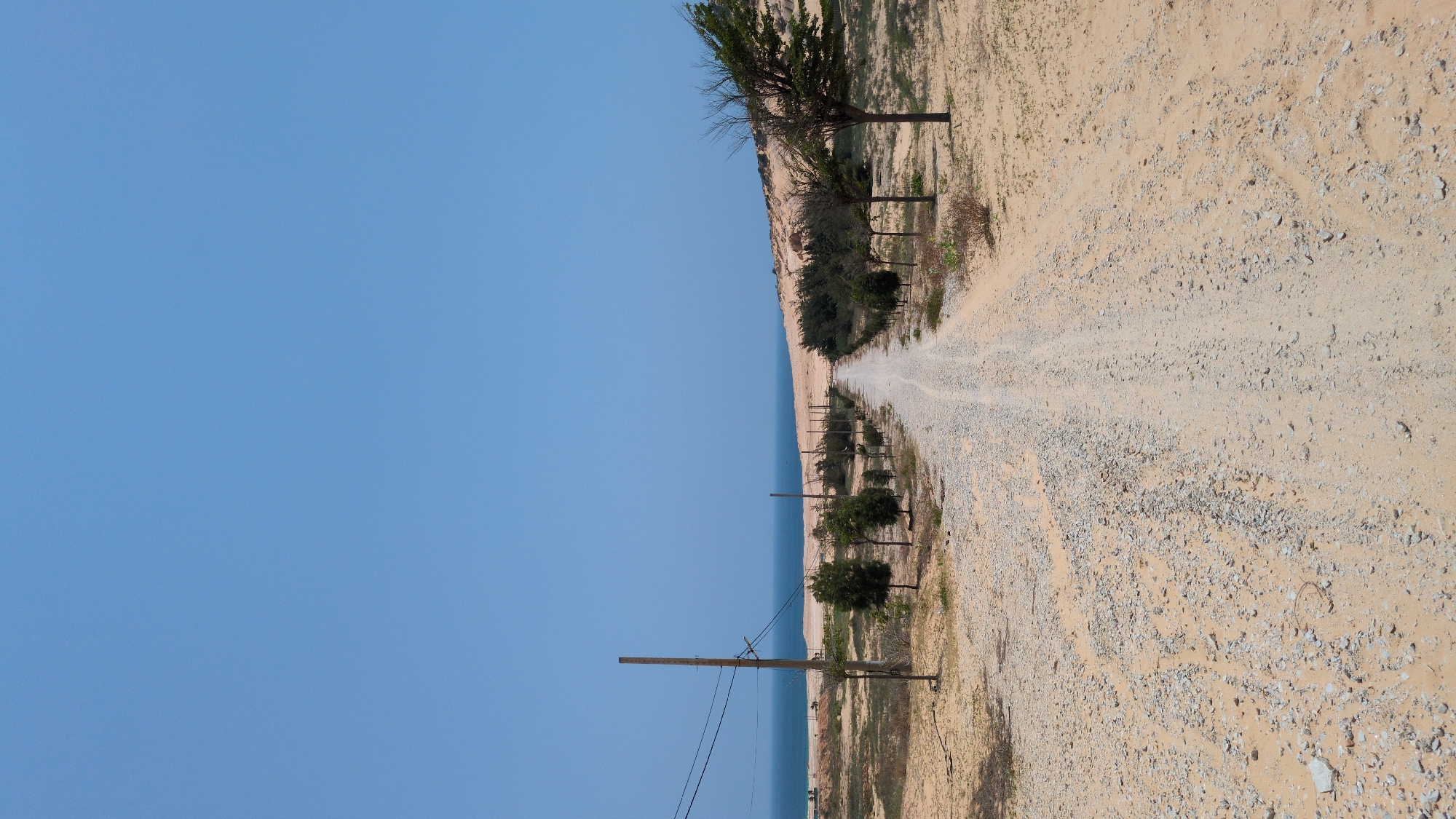 A ground-level, landscape-style photograph depicts a gravel road running parallel to the coastline. The scene is likely located near Phuoc Dinh, Vietnam. A clear blue sky dominates the left side of the frame. The ocean is visible as a blue stripe between the sky and the road. The road itself consists of sand and gravel, with tire tracks running along its length. The right side of the frame shows the landscape bordering the road, which is covered in sand, small rocks, and sparse vegetation. Trees and shrubs line the road, and utility poles with power lines are present. The lighting suggests a sunny day.