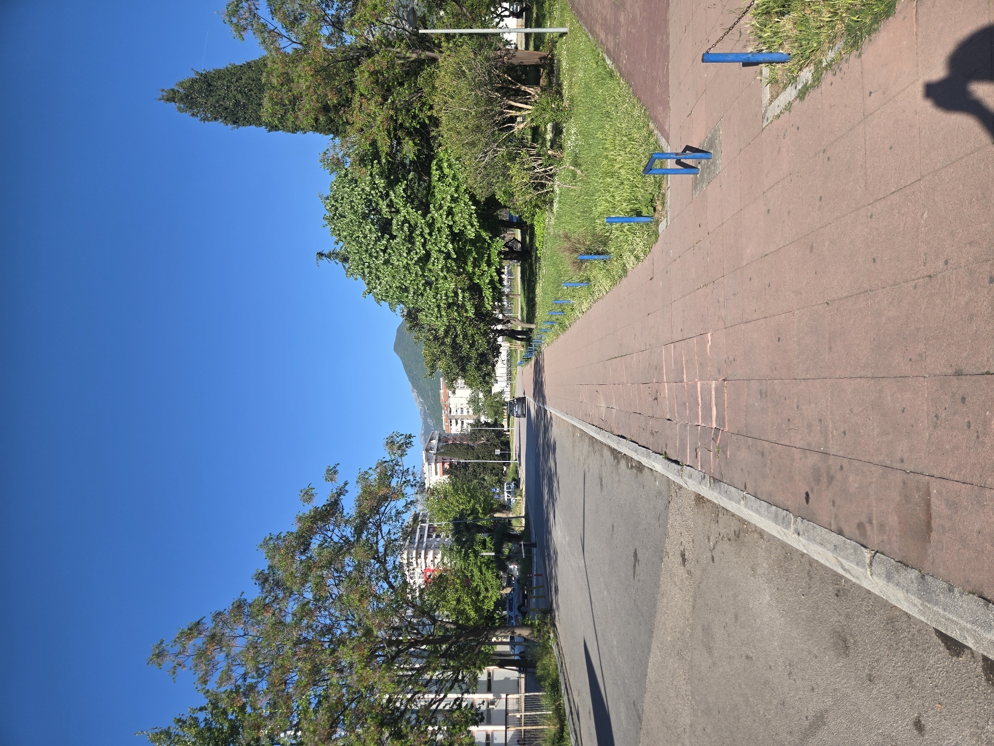 The image depicts a street scene in an urban area, likely in the city of Unknown, Unknown. The camera angle is low, emphasizing the perspective and depth of the scene. The sky is a clear, bright blue, contrasting with the greenery of the trees and the surrounding urban elements.
On the right side of the image, a paved walkway with reddish-brown tiles runs parallel to a gray asphalt road. Blue metal bars, possibly part of benches or barriers, are spaced along the walkway's edge where it meets a grassy area. The grass is a mix of green and yellow, indicating varying conditions.
The asphalt road appears to have been patched or repaired in places, showing an uneven surface. A curb separates the walkway from the road.
In the background, several buildings are visible, indicating an urban setting. Some trees line the street, providing shade and a sense of greenery. A mountain or large hill is partially visible in the distance, adding a natural element to the urban landscape.