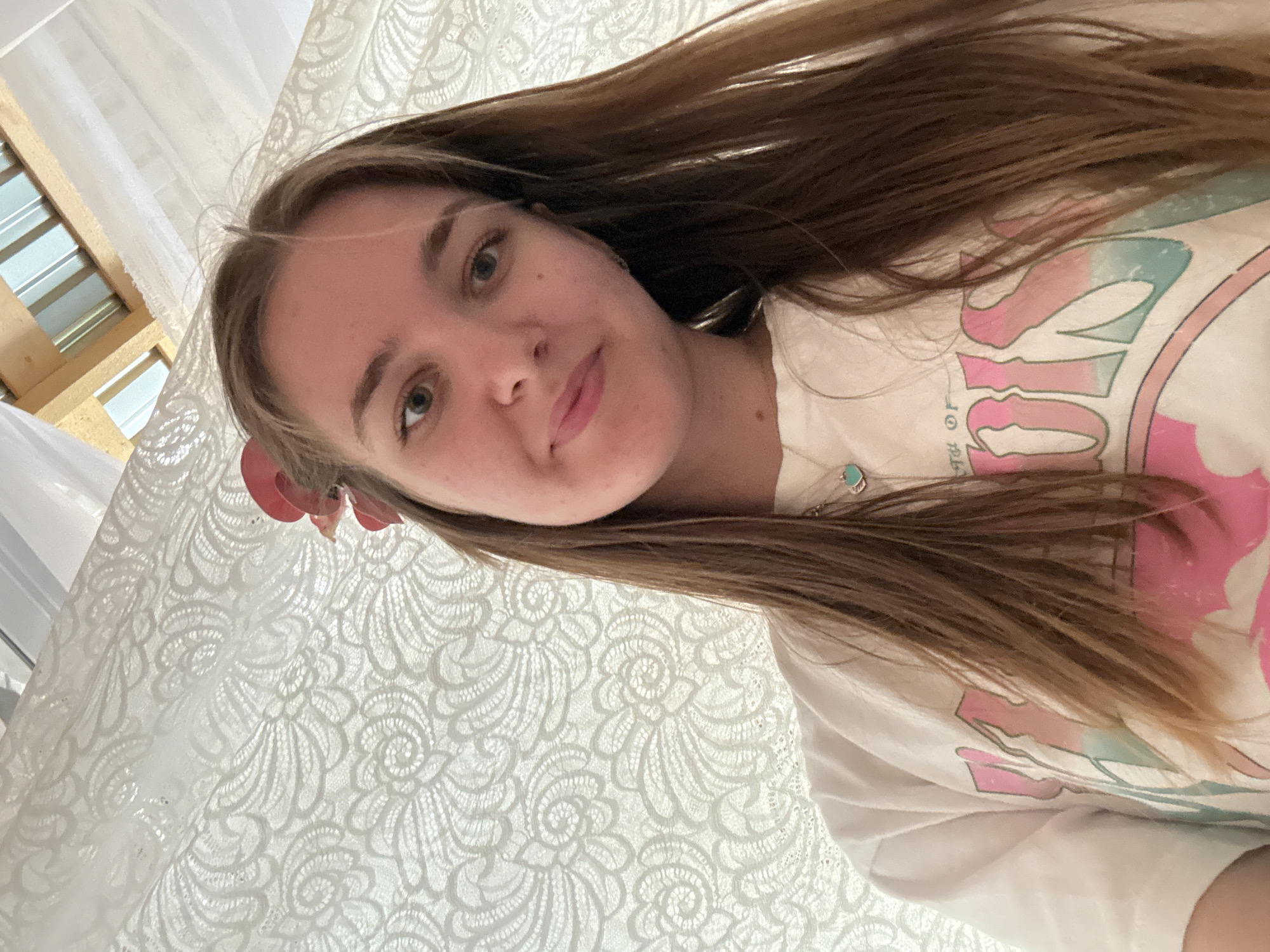 A young woman with light brown hair and fair skin takes a selfie at an elevated angle. She is wearing a white t-shirt with a pastel-colored graphic design and a necklace with a turquoise pendant. A red flower adorns her hair near her left ear. The background consists of a white lace fabric, possibly a curtain or tablecloth, and a window frame. The location is not recognizable as being in Sibir', Russia.