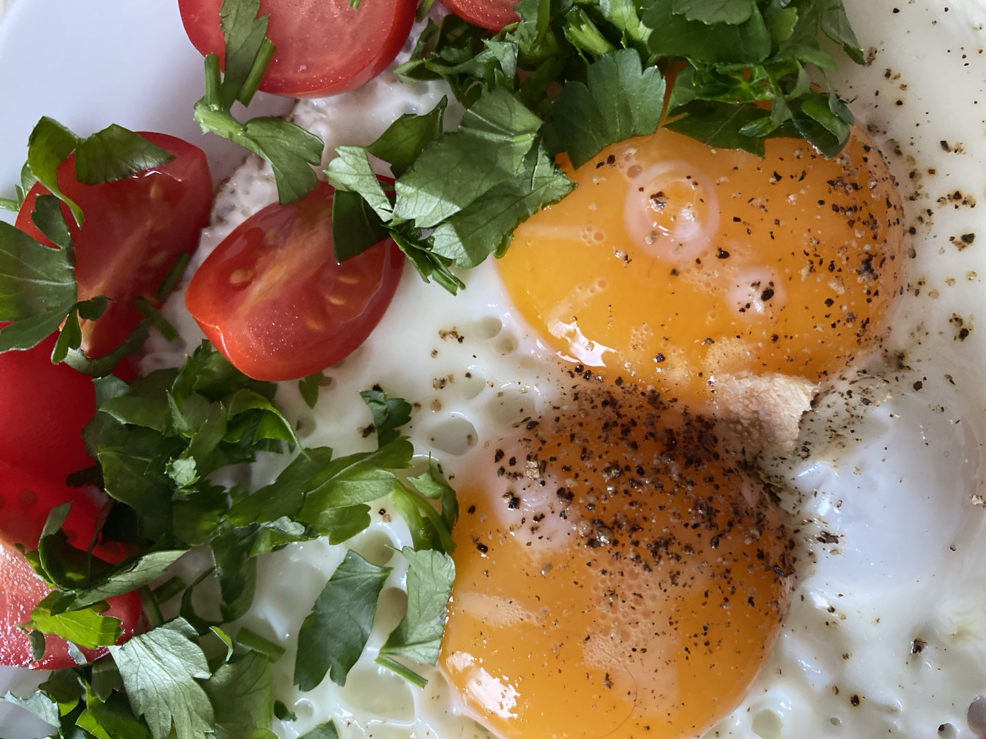 A close-up photograph shows two fried eggs seasoned with pepper, accompanied by quartered tomatoes and chopped parsley. The dish is presented on a white plate. There is no information that indicates the location in the city of Chaikovsky, Russia. There are no people visible in the image. The subjects are the fried eggs, tomatoes, and parsley. The object is the plate. The action is the preparation of food. There are no discernible interactions. The environment is a close-up food shot.