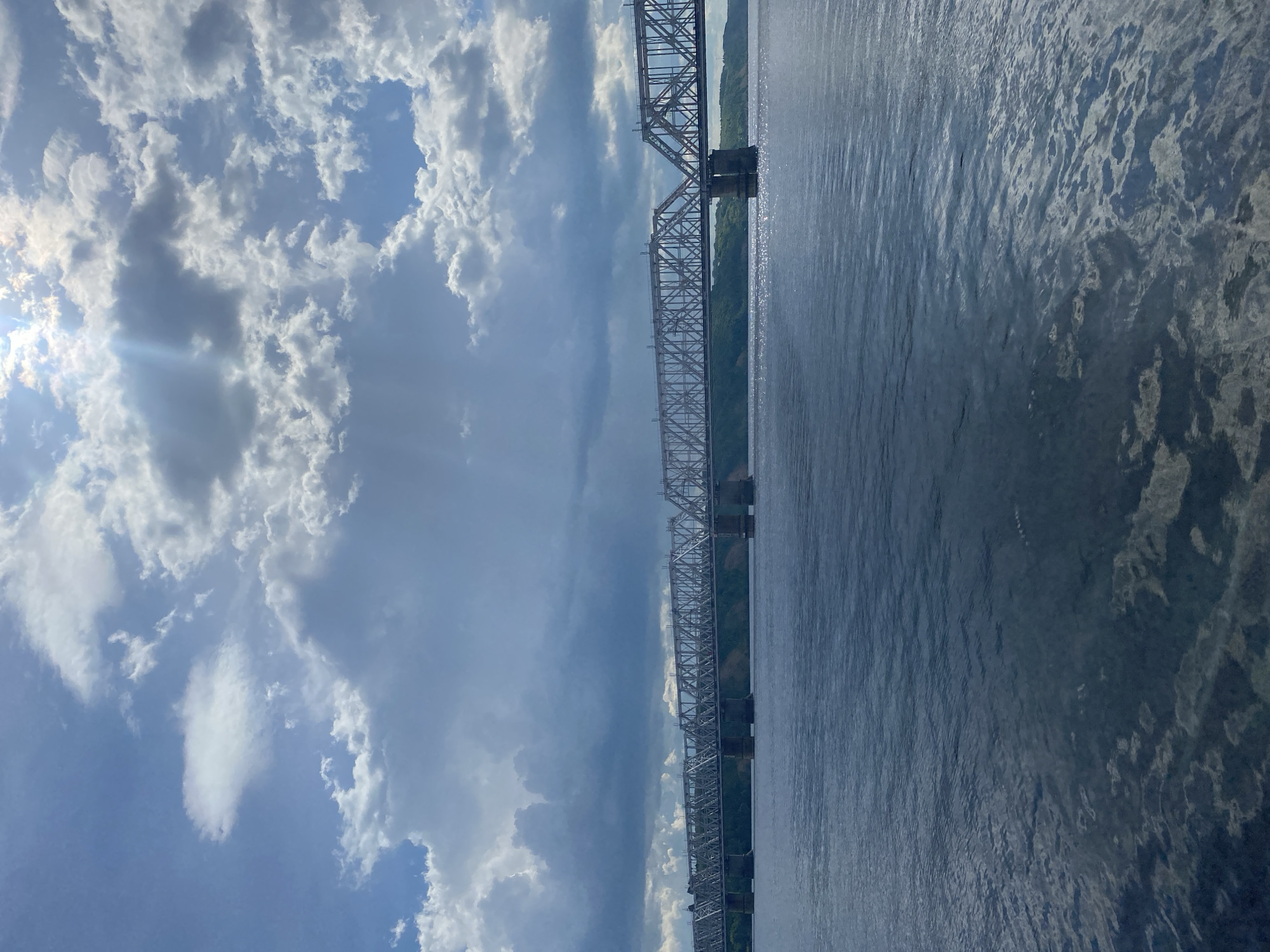 This media file depicts a wide, calm body of water alongside a large, steel truss bridge. The bridge spans a considerable distance, supported by multiple piers. On the left side of the frame, a dramatic sky is visible, filled with a mix of fluffy white clouds and darker, more ominous storm clouds. Sunlight breaks through in some areas, casting rays and creating lens flares. The water reflects the sky and the bridge, with subtle ripples disturbing the surface. Greenery lines the far bank of the water, partially obscured by the bridge's structure.
The scene appears to be captured during the daytime, given the presence of sunlight and the visible clouds. The weather seems dynamic, with both sunny patches and developing storm clouds. The presence of the large bridge suggests an industrialized or transportation-focused location. Without further information or visible text, a precise location cannot be determined, but the style of the bridge and the landscape could be consistent with a river setting. No people are visible in the media file. The overall mood is one of natural grandeur and perhaps a touch of atmospheric tension due to the weather.