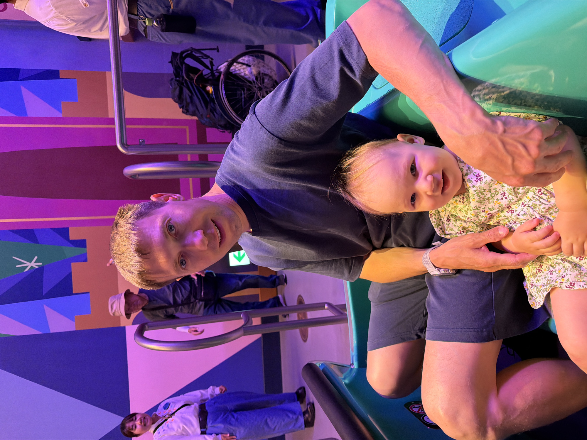 In a brightly lit indoor setting with walls adorned with abstract geometric shapes in shades of purple, blue, and green, a man and a baby are seated in a teal-colored vehicle. The man, with short blond hair and blue eyes, is wearing a dark t-shirt and dark shorts. He has a watch on his left wrist and is holding the baby with his right arm. The baby, facing the camera, has blonde hair and is wearing a floral-patterned dress.
In the background, other individuals are visible. To the left, a woman in a white shirt and blue pants is looking towards the camera and smiling. Behind her, a person wearing a hat and a dark shirt stands near a metal railing. Further back, a person in a white shirt and blue pants is also visible. A black backpack is placed on the ground near a bicycle wheel. A green arrow sign is mounted on the wall. The overall scene appears to be within a theme park, possibly Tokyo Disneyland, located in Urayasu, Japan, given the characteristic colorful and stylized environment.