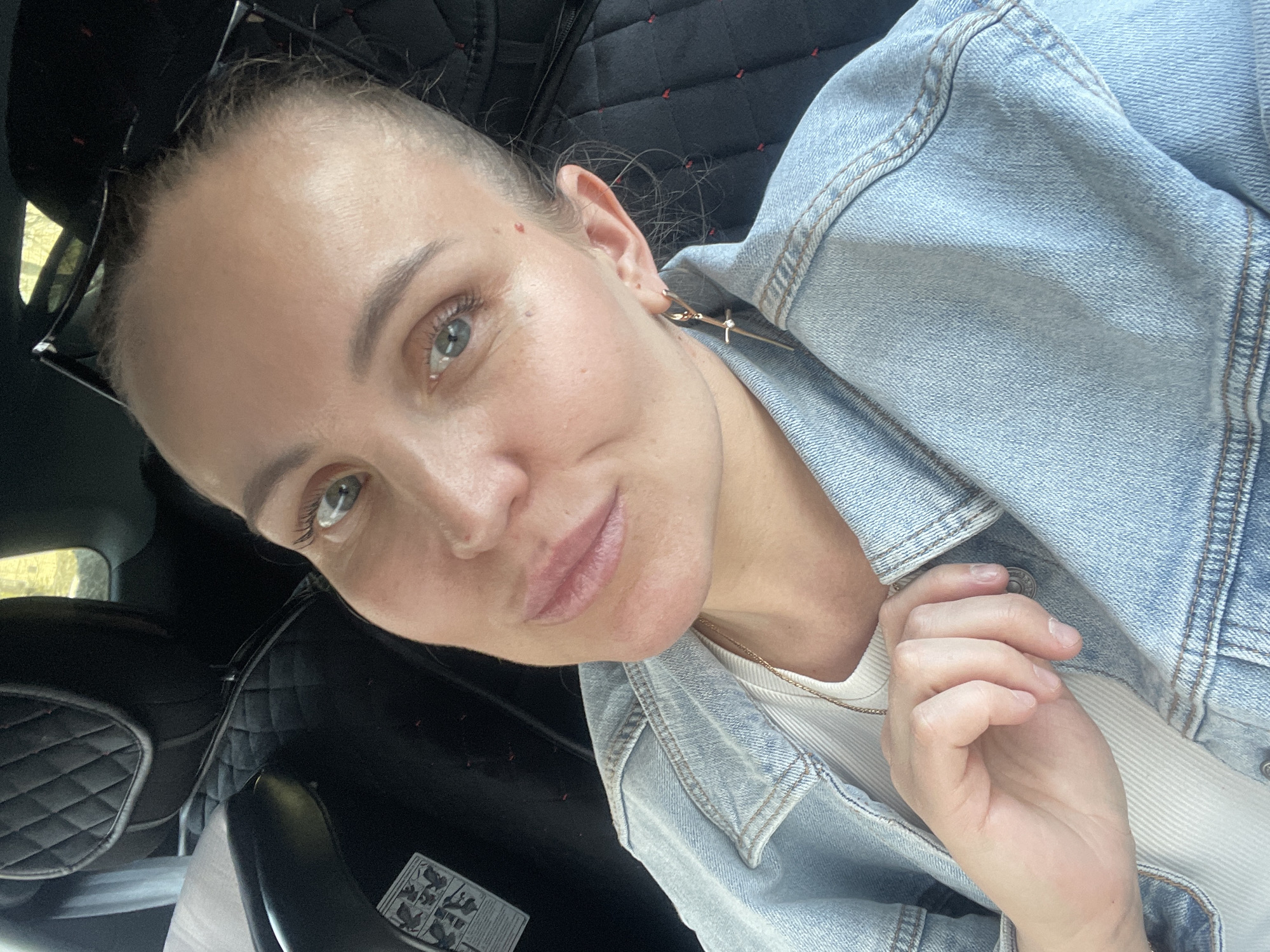 A woman is pictured from a low angle, her face and upper torso filling the frame. She has fair skin, light eyes, and her hair is pulled back tightly, exposing her forehead. She wears a light blue denim jacket over a white top. A thin gold necklace is visible at her neck. Her left hand is near her chest. The background shows a dark, quilted car seat with a safety instruction sticker. The photograph seems to be taken inside a car in the city of Chaikovsky, Russia. Natural light enters from a window on the left side of the frame.