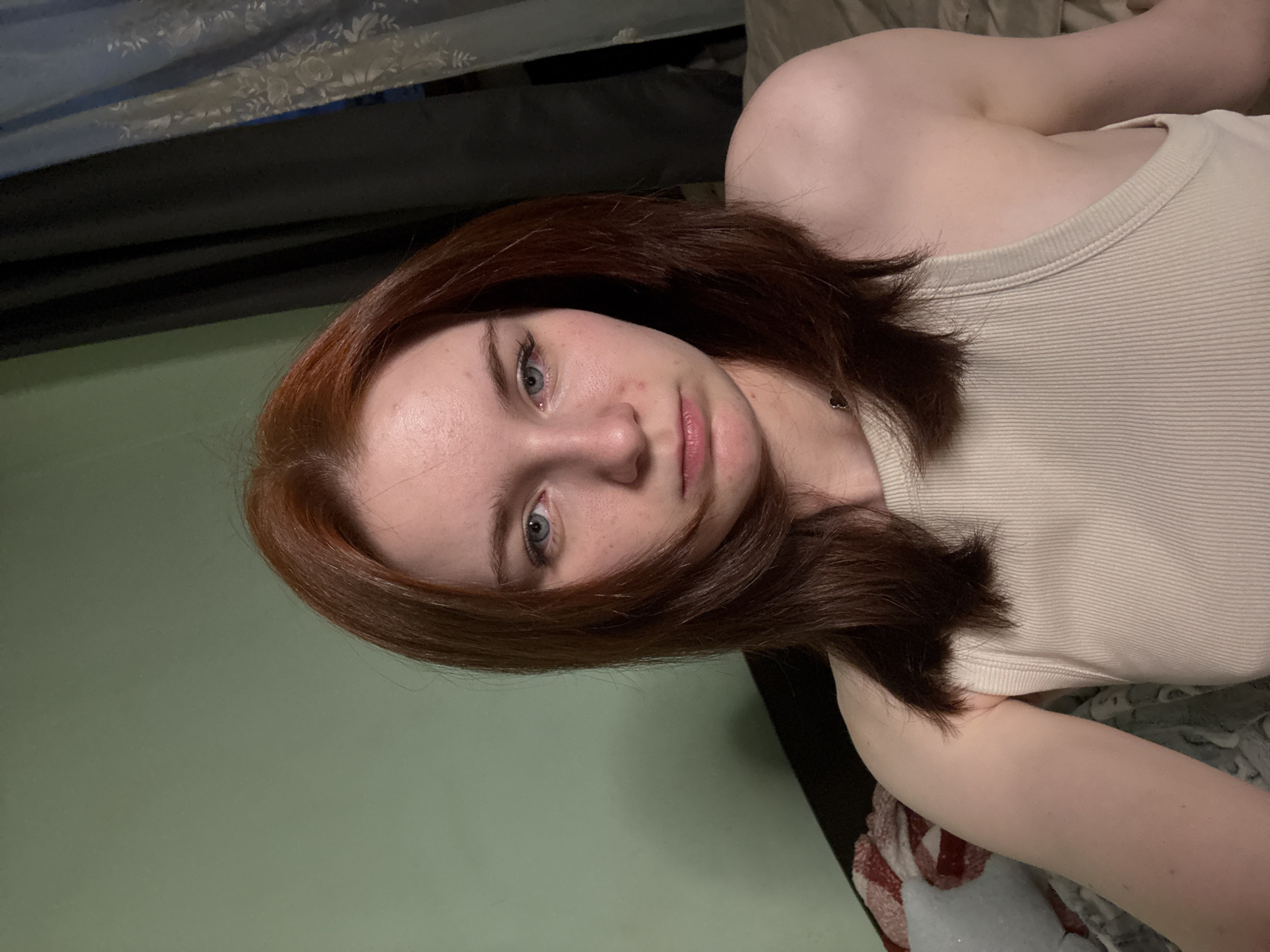 A close-up, angled shot depicts a fair-skinned young woman with auburn hair and blue eyes. She is wearing a ribbed, light-colored tank top. Her face is the primary focus. The background is a mix of green and darker, indeterminate elements. The setting appears to be indoors.