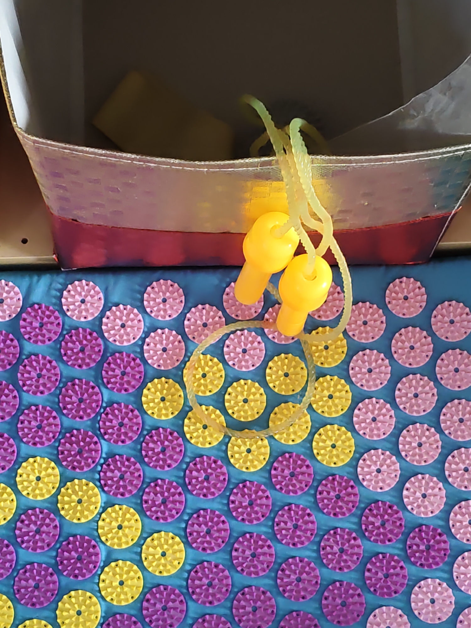 The image shows a close-up, top-down view of a turquoise acupressure mat with a pattern of purple, pink, and yellow circular plastic spikes. Draped over the mat are two yellow, earplug-like objects connected by a flexible, light green cord. Behind the mat, a silver and red decorative basket is partially visible, containing a yellow object, possibly a sponge. The scene is indoors, with no specific landmarks to indicate a location in Rostov-on-Don, Russia.