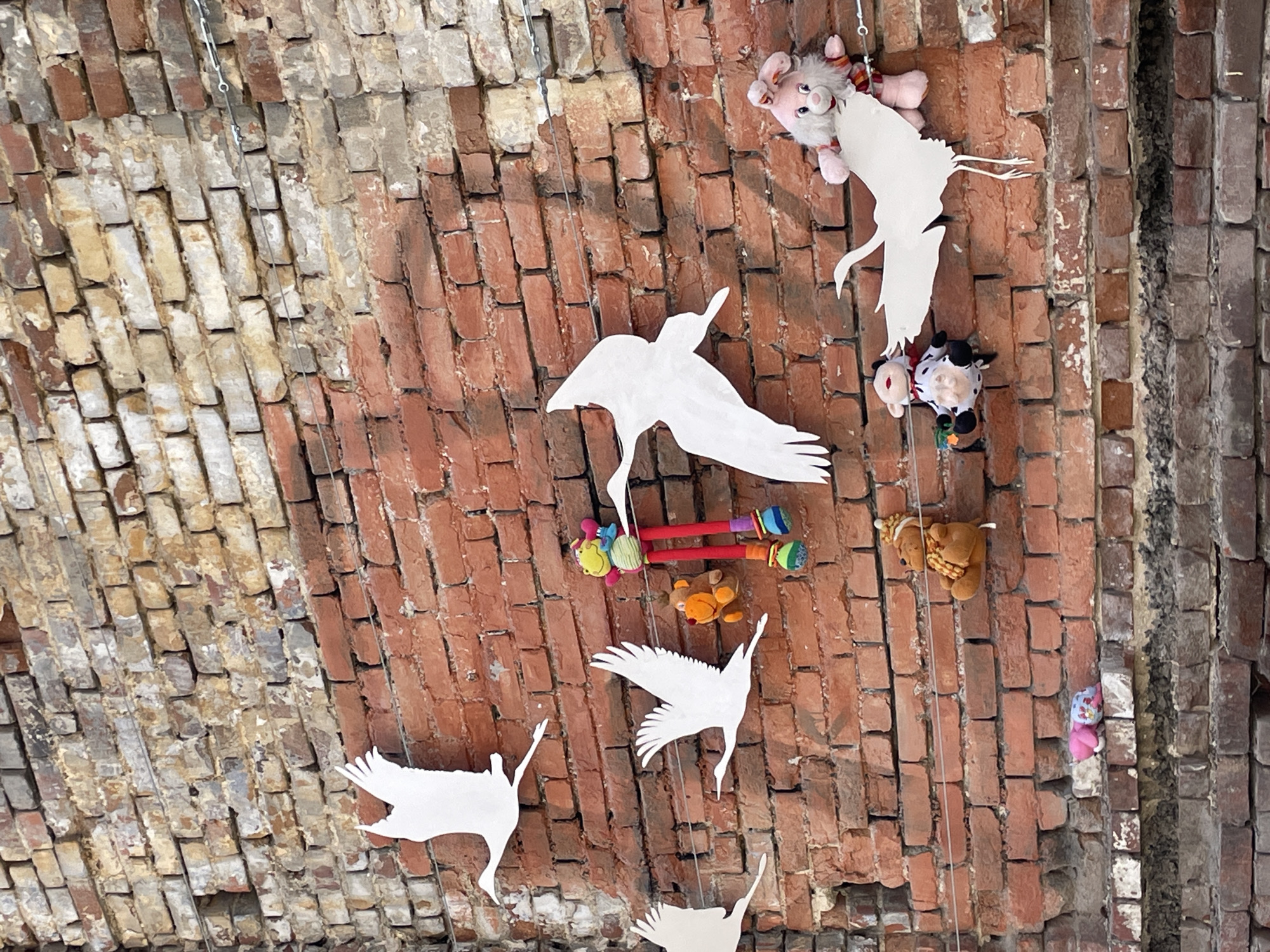 The image shows an upward view of a brick archway, possibly a ceiling or passageway, decorated with paper cutouts of flying storks and assorted stuffed animals. The brickwork is a mix of red and beige bricks, some showing signs of wear and discoloration. The paper storks are white and stylized, suspended at various points along the arch. A variety of stuffed animals, including a pink rabbit, a cow, a teddy bear, and colorful fabric dolls, are also suspended from the archway. The scene appears to be in a public space, possibly an art installation or children's area. This location is in the city of Yekaterinburg, Russia.
