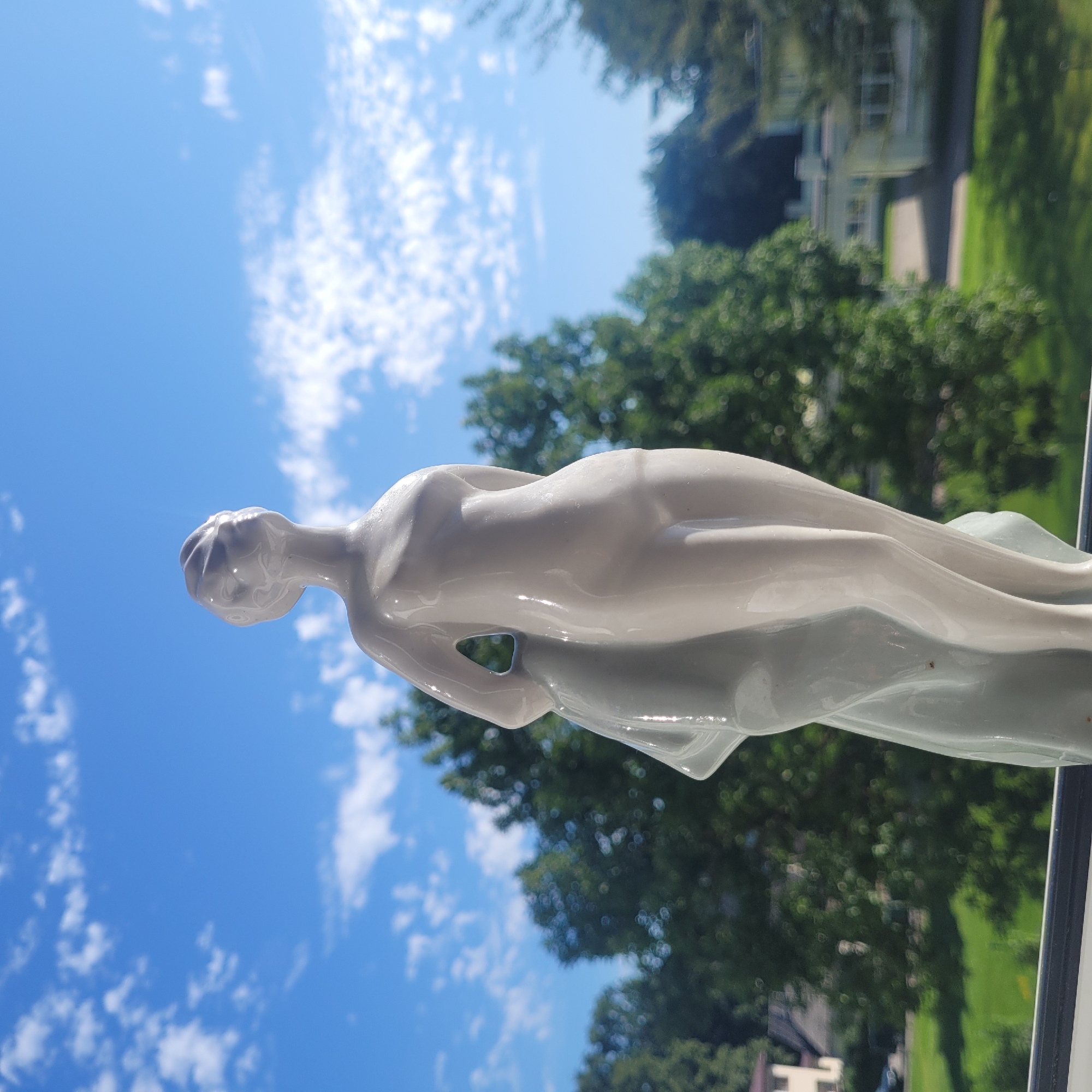 A white ceramic figurine of a woman stands against a bright blue sky with scattered white clouds. The figurine is positioned in the foreground, with its gaze directed upward and to the right. A flowing white fabric drapes around the figure's left side. The background consists of lush green trees and a portion of a building, suggesting an outdoor setting in a suburban or park-like area. The lighting indicates daytime, with sunlight illuminating the figurine and creating subtle shadows. The image appears to have been taken from a slightly low angle, looking up at the figurine.