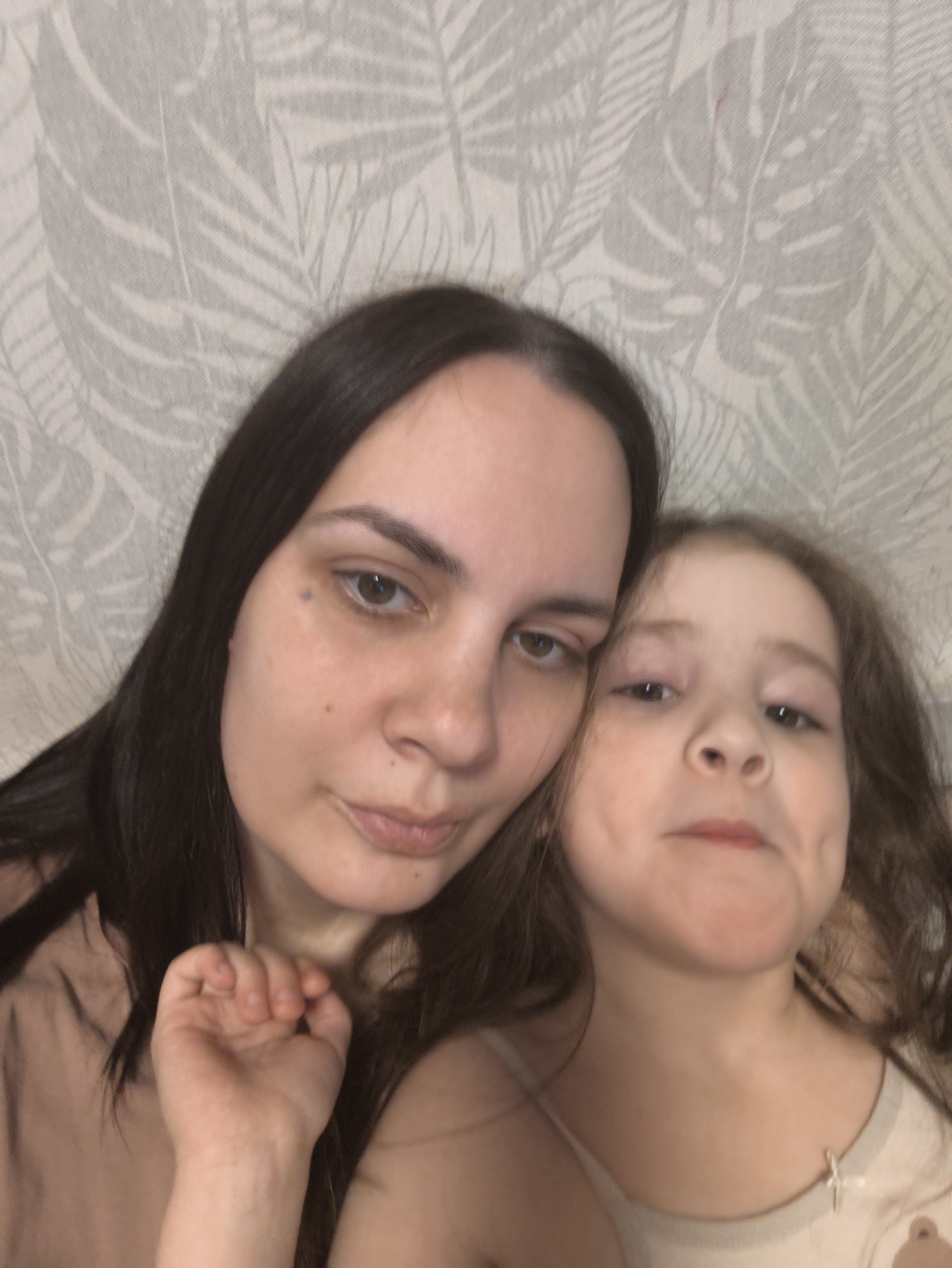 A selfie shows a woman and a young girl. The woman has dark hair, light skin, and is positioned on the left. Her hand is raised near her face. The girl, on the right, has brown curly hair and light skin. Both are looking towards the camera. The background is a wall with a light-colored leafy pattern. The location appears to be indoors. There is no information in the image to connect it to Chaikovsky, Russia.