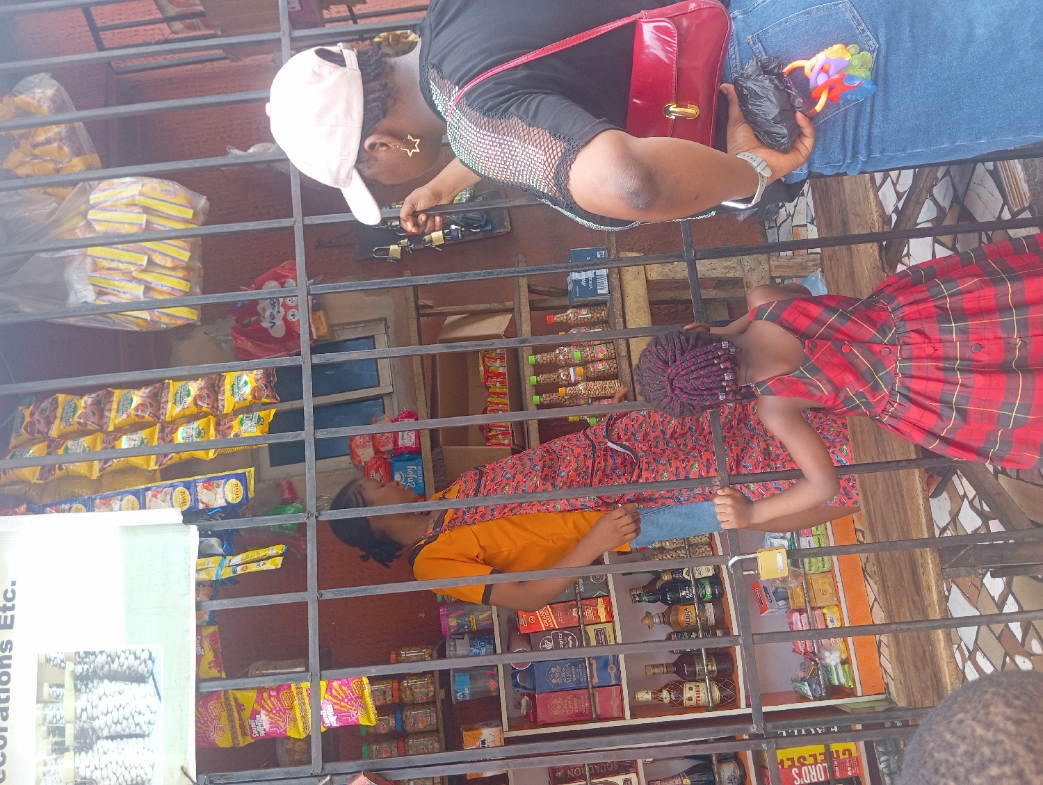 The image, rotated counter-clockwise, captures an everyday scene at a street-side shop in Nkpor, Nigeria, viewed through security bars.
In the foreground, an adult woman wearing a black top, blue jeans, and a white baseball cap stands. A red handbag is slung over her shoulder. She holds a small black plastic bag and a colorful child's toy or rattle in her left hand, while her right hand is near some hanging keys and padlocks inside the shop. She appears to be either making a purchase or browsing items. Visible beneath her cap are dreadlocks, and she wears star-shaped earrings.
To her right, a young girl in a red and black plaid dress stands next to the security bars, looking into the shop. She has red braided hair.
Behind the security bars, inside the shop, another person (likely the shopkeeper) wearing an orange top and a patterned skirt or wrapper is partially visible, attending to the goods.
The shop is well-stocked with various consumer goods displayed on shelves. These include numerous packets of snacks and biscuits (many in yellow packaging), bottles of drinks (some appearing to be spirits or liqueurs, others non-alcoholic beverages), and packaged toiletries such as diapers or sanitary pads. A white sign on the far left partially reads "Decorations Etc." On a lower shelf, a bottle label partially shows "LORD'S" and "GRE" (likely "GREEN"). The scene depicts a typical daytime transaction at a local convenience or provision store. No specific weather cues are visible, but the bright lighting suggests daytime.