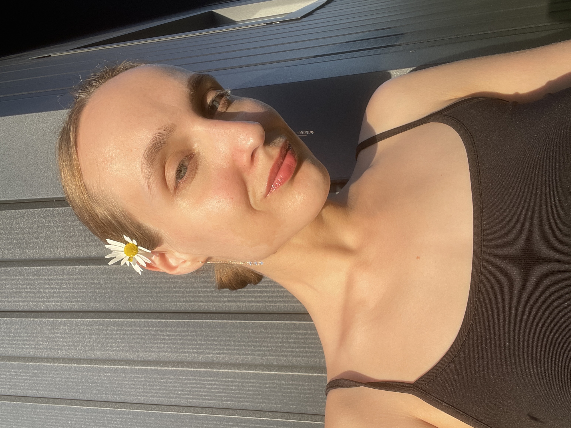 A woman with fair skin, blonde hair, and light-colored eyes is positioned at an angle against a gray, horizontally-lined surface. She is wearing a dark tank top. A daisy is tucked behind her ear, and a chain earring with small, clear beads dangles from her other ear. The lighting is bright, with a notable contrast of light and shadow across her face and the background. The location is given as Подшивалово, Russia, but there is no visual evidence in the image to verify this.